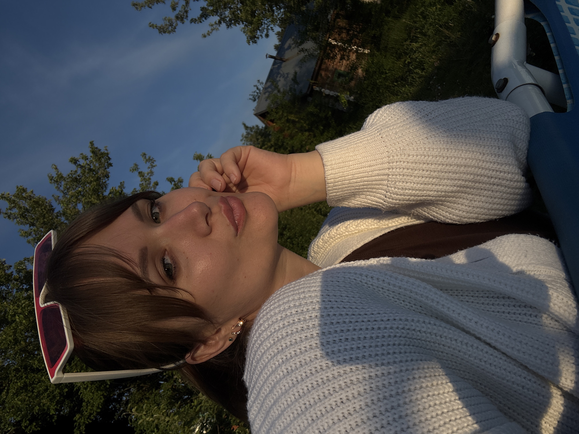A young woman is depicted in a close-up, tilted portrait, appearing to be reclining or leaning back outdoors. She has dark brown hair with bangs, and pink and white cat-eye sunglasses are perched on top of her head. Her light-colored eyes (possibly green or hazel) look directly at the viewer, and her lips are a soft pink. She wears a thick, white knit sweater over a dark brown garment. Her right hand is gently raised, with fingers resting near her chin and jawline. In her left ear, two small gold earrings are visible: one a spiral ear cuff and the other a stud.
The background features a clear blue sky with some wispy clouds, and lush green trees fill parts of the frame. Through the trees, a rustic-looking wooden building with a dark roof and chimney can be seen. Below her right arm, a white and blue metal structure, possibly part of outdoor furniture like a swing or bench, is visible. The lighting is warm and soft, indicative of the golden hour, likely late afternoon or early evening, casting gentle highlights on her face and sweater and creating some depth with shadows. The overall scene suggests a tranquil outdoor setting in Chaikovsky, Russia. There is no visible text in the image.