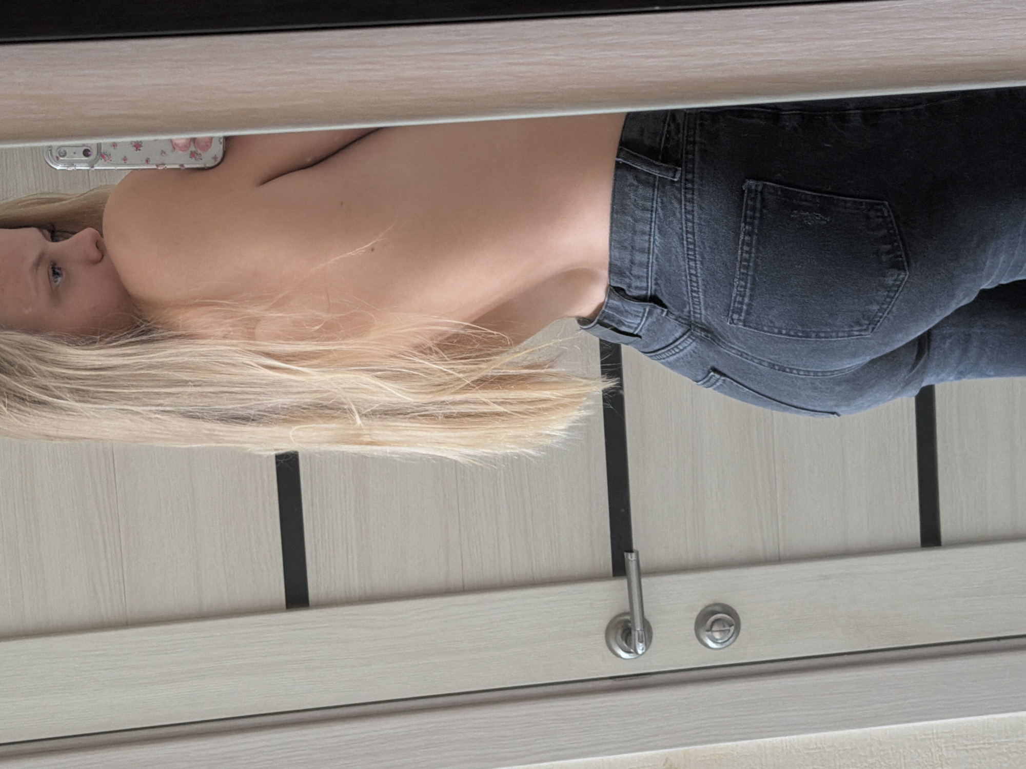The image shows a person from the waist up, wearing dark jeans and partially bare-backed, holding a phone to a mirror. The person has blonde hair. The background is a light-colored door with vertical, dark-colored inlays and a silver-colored handle. No information about Izhevsk, Russia is discernible from the image.
