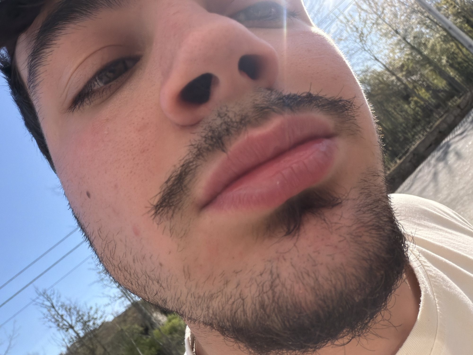 A close-up, slightly angled selfie captures a person's face. The subject has dark hair and a dark beard. The person appears to be outdoors. Power lines and trees are visible in the background suggesting an urban or suburban location. The person is wearing a light-colored shirt. Based on the limited visual information, a precise location in Vila Real, Portugal, cannot be determined.