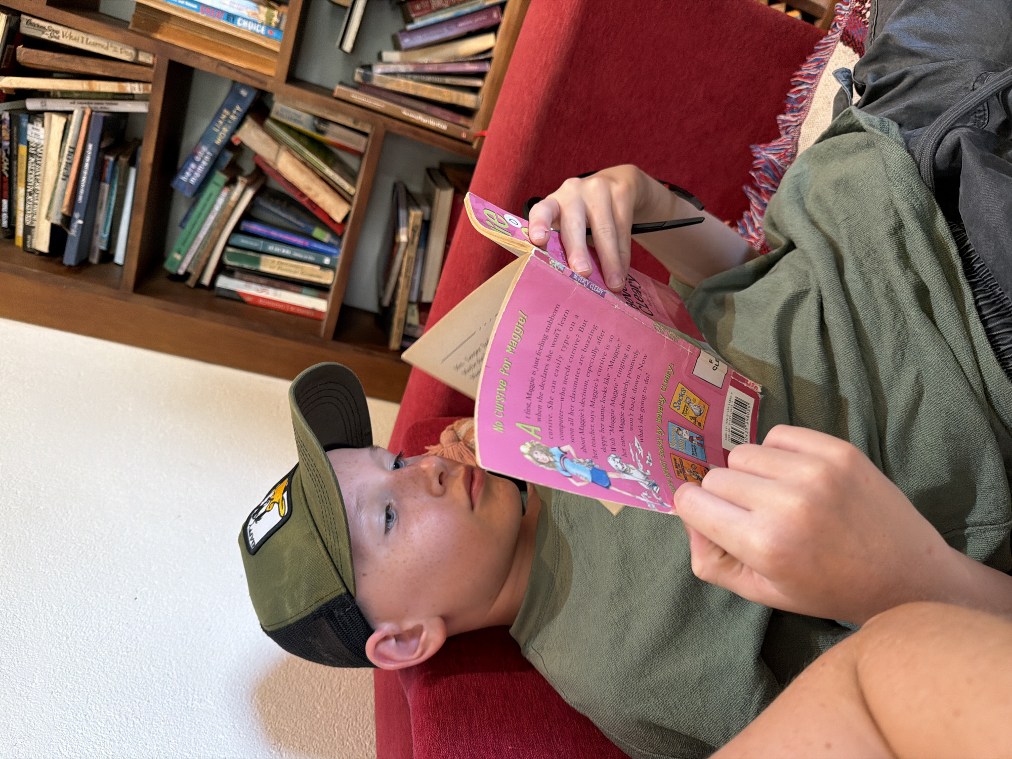 An undated, medium shot, eye-level, outdoor photograph shows a Caucasian boy reading a pink book. The boy is wearing a green t-shirt, a green and black baseball cap, and gray pants. He is sitting on a red chair. Behind him, there is a bookshelf filled with books. The boy's left arm and hand are visible, holding the book. The background is blurred, but it appears to be a room with a bookshelf and a wall. The lighting is natural. The photograph does not provide any information about the location in the city of Đà Nẵng, Vietnam.