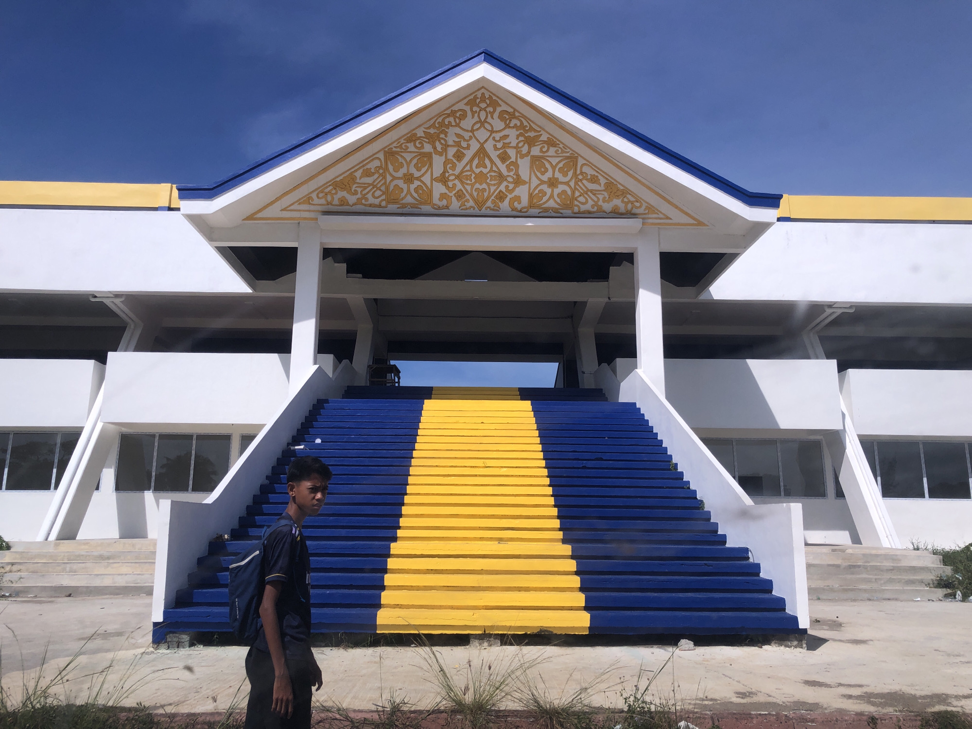 A young person wearing a dark shirt and a backpack stands in front of a large building in Tambontunong, Indonesia. The building is primarily white, with blue and yellow accents. Prominent stairs with alternating blue and yellow steps lead up to a covered entrance adorned with a gold-colored decorative design. The sky above is clear and blue. Sparse vegetation grows in the foreground.
