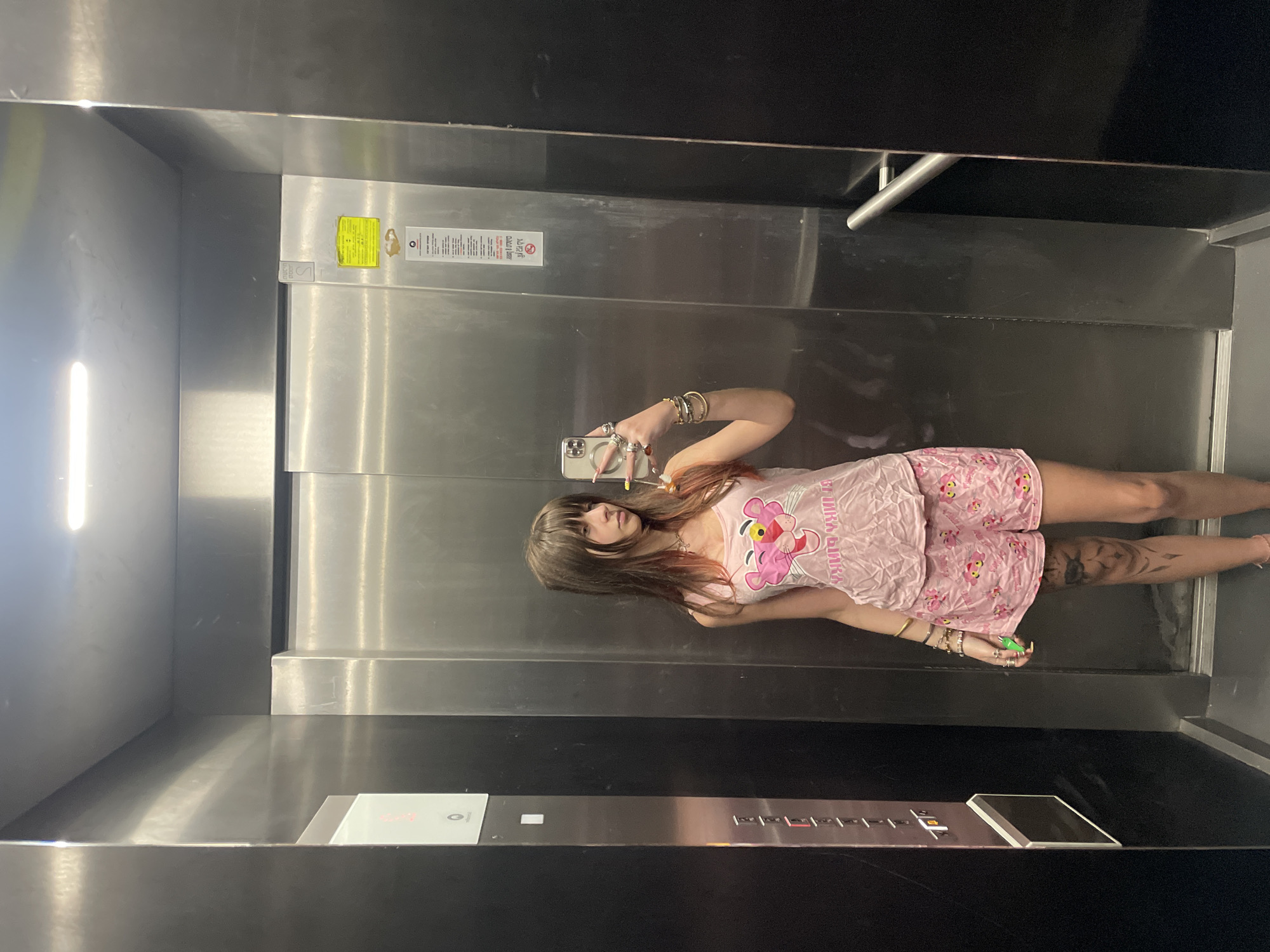 A person stands in an elevator, taking a selfie in the mirror. The elevator has stainless steel walls and a black ceiling. A yellow warning sticker is visible on one of the walls. The elevator's control panel is situated on the lower right side of the mirror, featuring several buttons. The person in the image is wearing pink pajamas with the Pink Panther cartoon character. They hold a smartphone to take the photo. No specific identifying information relates this elevator to Raanana, Israel.