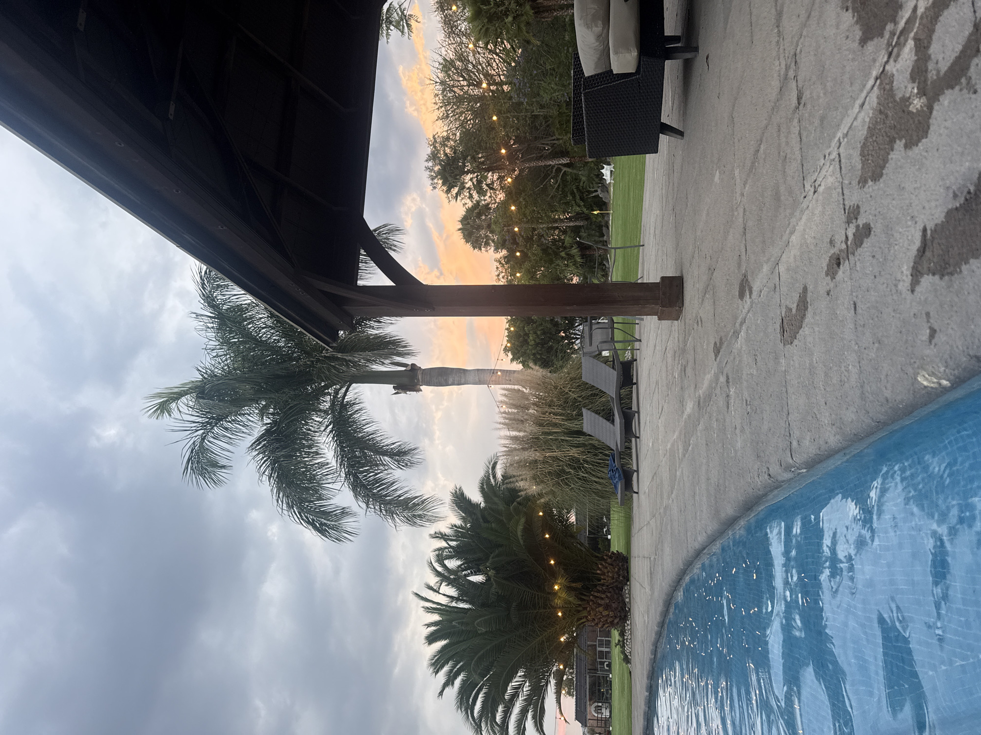 The image shows an outdoor scene at dusk or dawn in Santa Ana Yancuitlalpan (Chilhuacán), Mexico. The view is from under a dark, wooden structure, possibly a gazebo or pergola. A tall palm tree extends upwards toward a sky with a mix of grey clouds and a strip of orange-tinted sky, suggesting either sunrise or sunset. A second, shorter palm tree is decorated with string lights. To the right, a patio area is visible, with outdoor furniture including chairs and cushions. In the foreground, the edge of a blue-tiled swimming pool is visible. In the background, additional trees are strung with lights. The patio area appears wet, suggesting recent rain. There are no people visible in the scene.