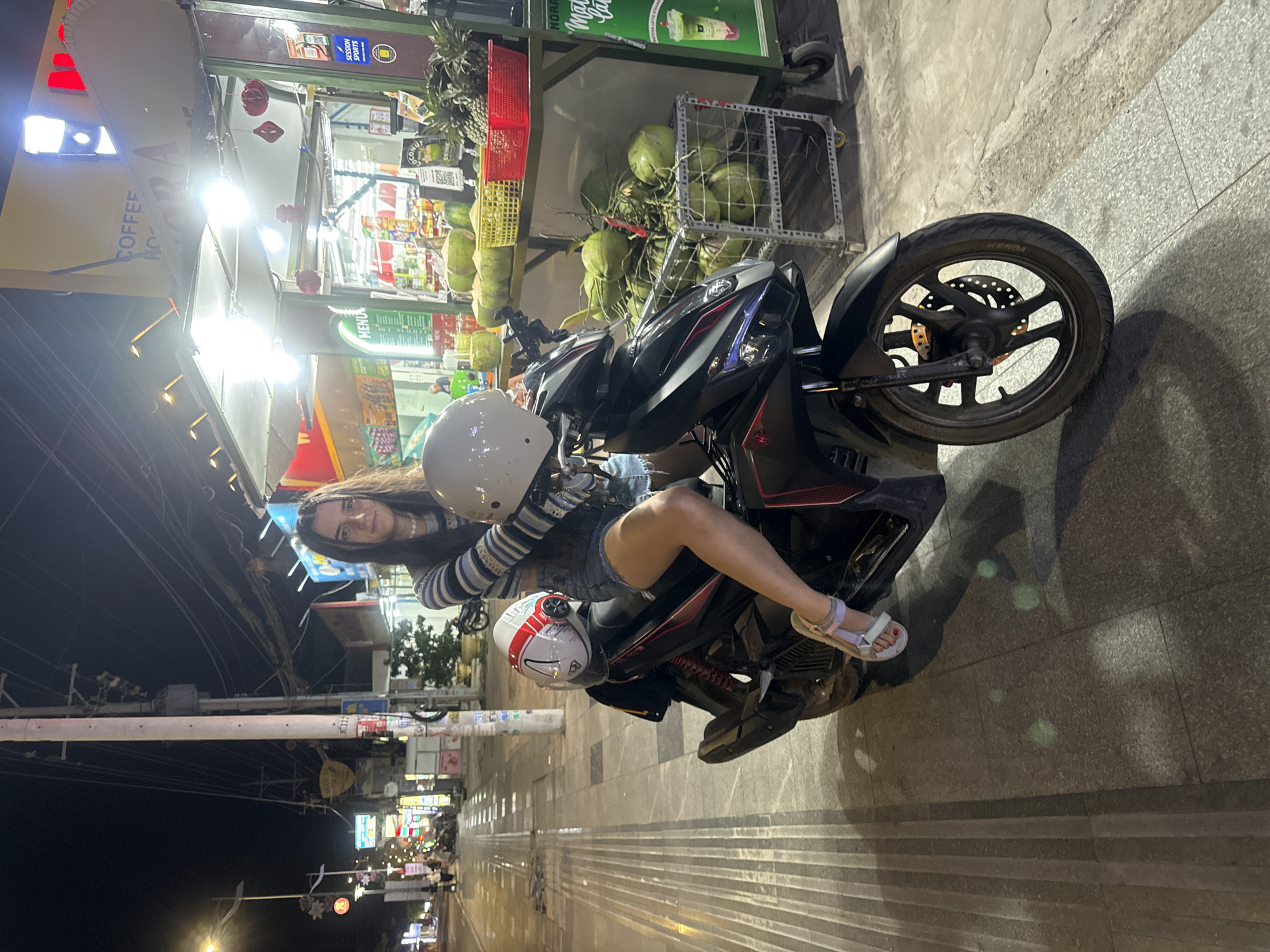 This image captures a vibrant nighttime street scene in Mũi Né, Vietnam, featuring a young woman posing on a scooter next to a brightly lit fruit and drink stall. The woman, with long dark hair, is seated on the black scooter, wearing a striped long-sleeve top and denim shorts, and has a slight smile directed at the camera. A white helmet rests on the scooter's handle, and another white helmet with red accents is visible on the seat behind her.
To the right of the scooter, a lively street vendor stall glows with bright lights, showcasing an abundance of tropical fruits like green coconuts and pineapples in metal and plastic baskets. Various packaged drinks and bottles are also visible inside the stall. Signs above the stall display text including "COFFEE," "ROMA," "MENU," "SESSION SPORTS," and "COCONUT," along with QR codes, suggesting a place where locals and tourists can buy refreshments.
The setting is an urban street or sidewalk, paved with grey tiles. In the background to the left, the darkened sky is contrasted by the distant glow of streetlights and businesses further down the road, with utility poles and tangled wires typical of Southeast Asian infrastructure. The scooter's rear tire prominently shows the brand "KENDA." The overall atmosphere is relaxed and typical of an evening in a bustling Vietnamese town.