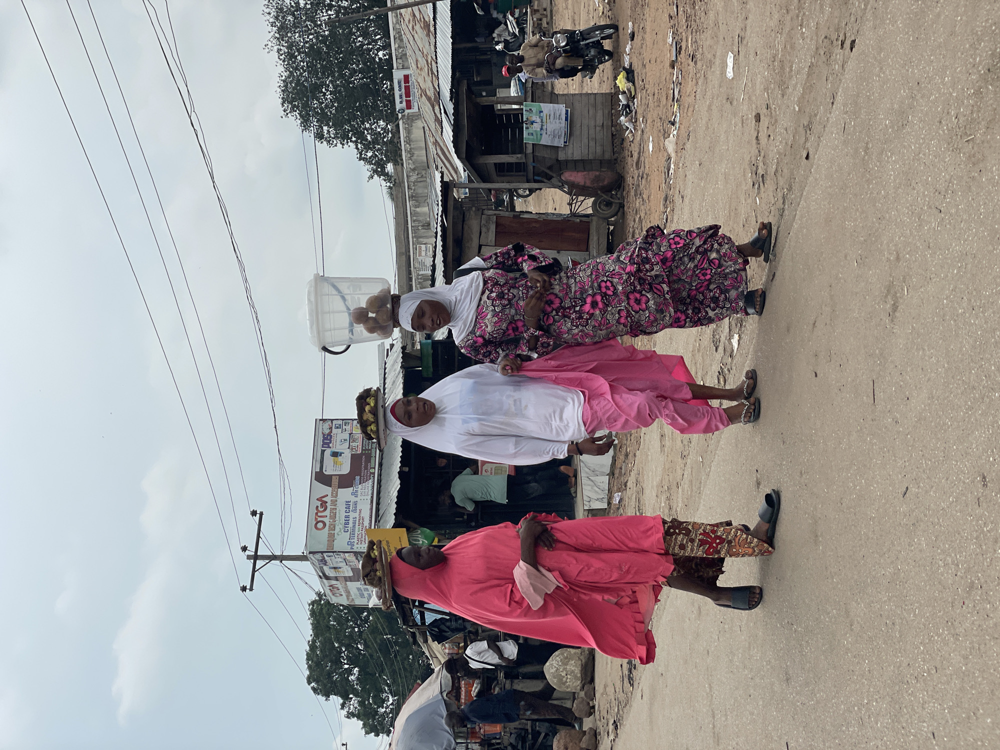 In Lokoja, Nigeria, three women walk along a dusty street lined with low-rise buildings and stalls. The woman on the left wears a bright pink abaya and headscarf, carrying a flat circular object piled with food items on her head, balanced by her hands. She wears black slip-on sandals and patterned trousers peeking out from under her abaya. To her right, a woman in a white hijab and pink pants also carries a similar load on her head. She wears sandals. The woman on the far right is dressed in a vibrant pink and black floral patterned abaya and white hijab, and she also carries a large clear plastic bucket filled with round, brown food items on her head. She wears black slip-on sandals. In the background, other individuals are visible, some tending to stalls, and a man sits on a motorcycle. The environment appears to be a busy market area with overhead electrical wires crisscrossing the sky. Debris is scattered on the ground.