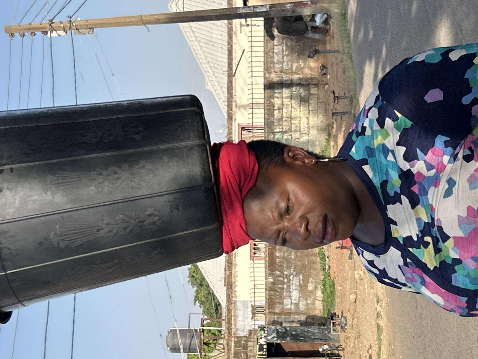 An African woman balances a black, patterned water tank on her head, secured by a red cloth, on a street in Jos, Nigeria. She wears a floral-patterned blue, white, and pink shirt. In the background are buildings, a water tower, an electricity pole, and greenery. The street is paved and appears to be in a residential area.