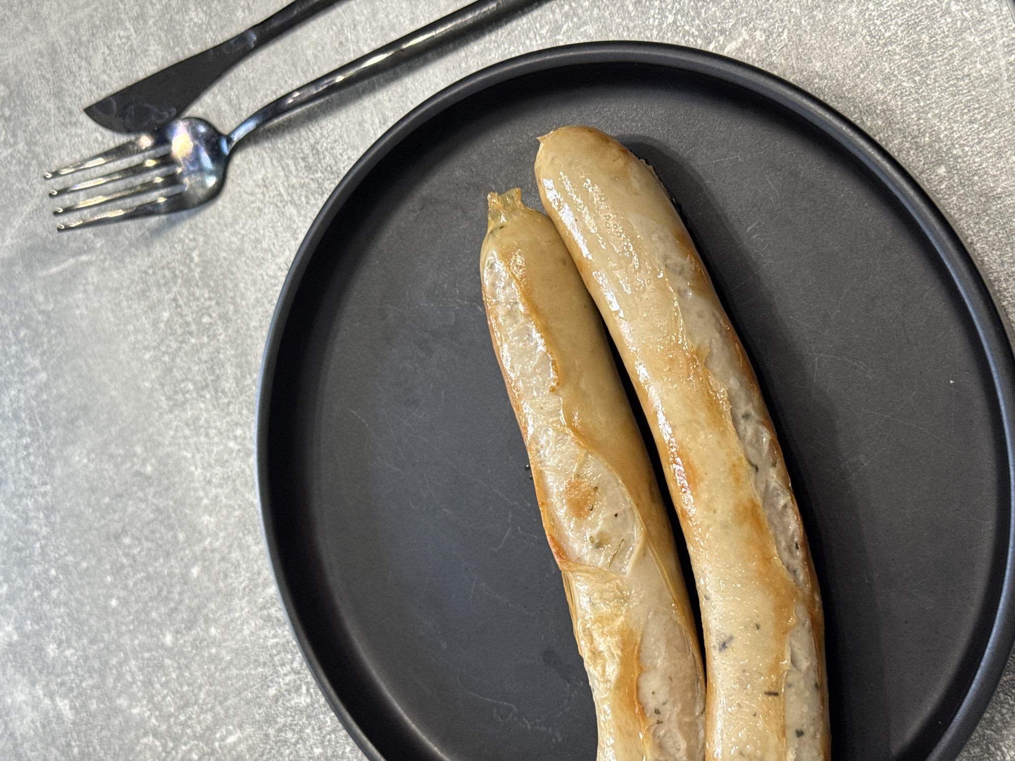 Two cooked sausages are presented on a black circular plate, which rests on a textured gray surface that resembles a countertop or table. To the upper right of the plate, a silver fork and a black knife are arranged, suggesting a meal setting. The sausages are golden brown with some charring and appear to be seasoned with herbs. The lighting is bright, casting a slight sheen on the sausages and highlighting the texture of the surface. There are no people visible in the image, and no specific landmarks or clues are present to indicate a location in Lyubertsy, Russia.