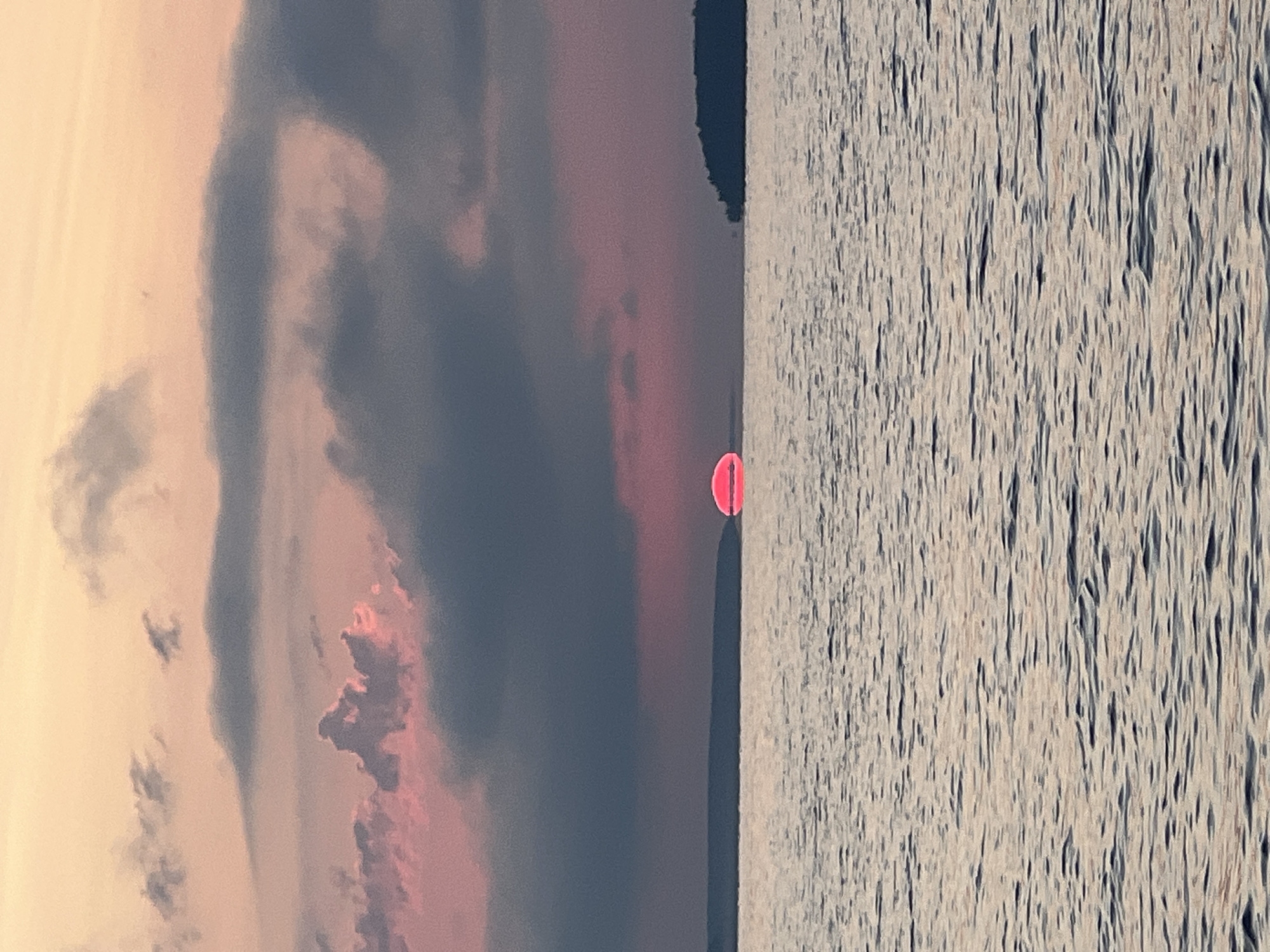 A sunset over a body of water in Novopichugovo, Russia, features a bright pink sun partially obscured by dark, textured clouds. The sun sits low on the horizon, casting a reddish glow on the water's surface. Two silhouetted landmasses, one long and thin and another a more distinct island shape, frame the scene on the left and right respectively. The water is rippled, reflecting the muted colors of the sky. The overall impression is one of a serene and colorful end to the day.