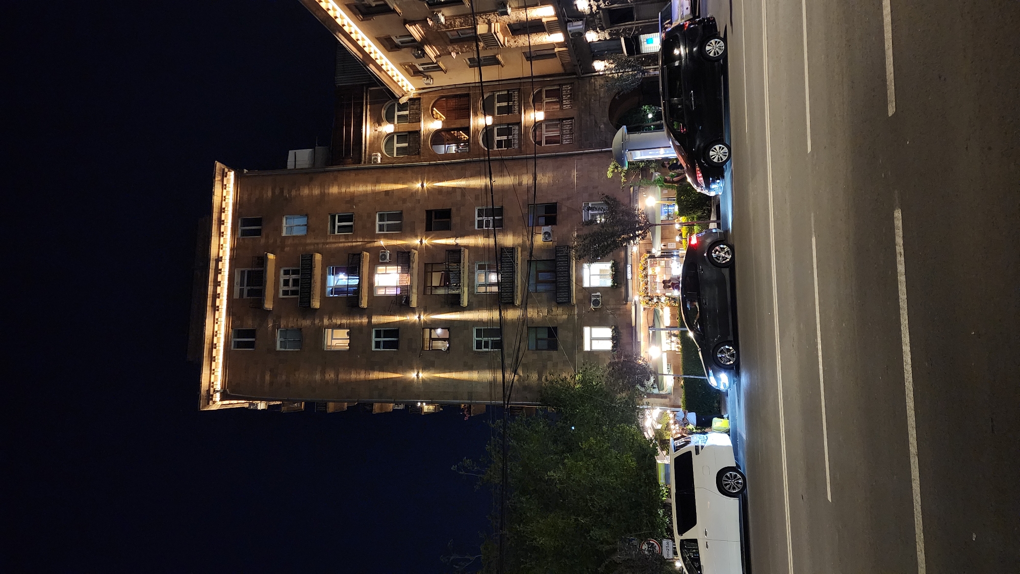 This is a nighttime photograph taken in Yerevan, Armenia, featuring a multi-story building and a street scene. The building is constructed of light-colored stone and is illuminated by decorative lights along its facade, highlighting the windows and architectural details. Some windows are lit from within, suggesting occupied apartments or businesses. Below the building, trees and greenery are visible, adding a touch of nature to the urban environment.
The street in front of the building is paved and has white lane markings, indicating it is a public road. Two dark-colored cars and one white van are parked along the street. The vehicles are illuminated by streetlights and their own headlights and taillights, creating reflections on the wet-looking pavement. The overall atmosphere is one of a quiet urban evening. The time of day is clearly night, and the presence of artificial lighting suggests an absence of natural daylight. There are no people prominently visible in the frame. No text is legible in the image.