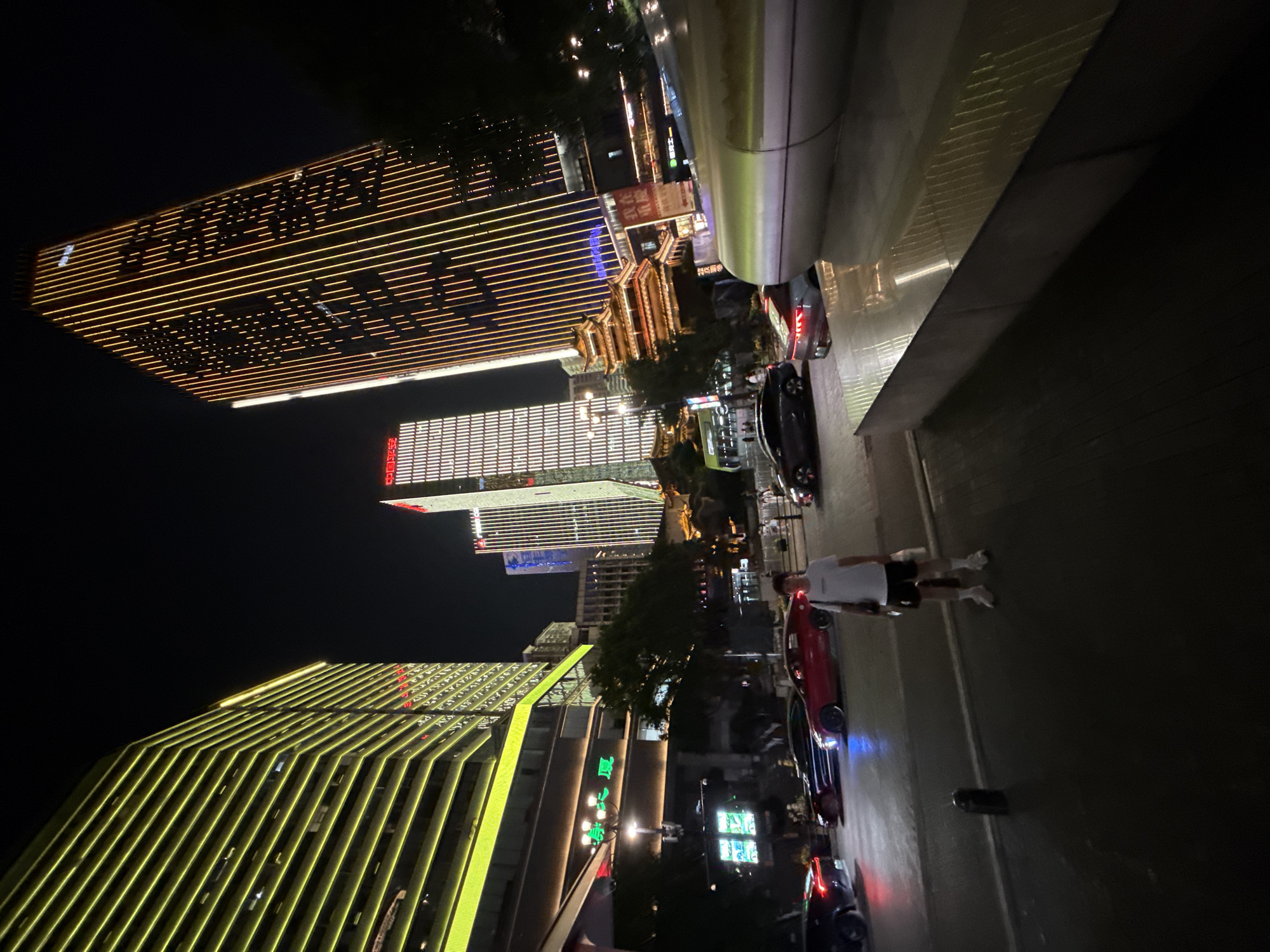 The image depicts a nocturnal urban scene in Yuzhong District, China, characterized by illuminated skyscrapers and street-level activity. Dominating the background are several modern high-rise buildings, some adorned with vertical yellow and white LED lights forming intricate patterns, while others feature less elaborate lighting. In the mid-ground, a traditional Chinese-style pagoda-like structure is nestled among the modern architecture. A street runs horizontally across the scene, populated by several vehicles including a red sports car, a dark sedan, and other cars partially visible. On the sidewalk in the foreground, a young person wearing a white t-shirt and dark shorts stands facing the camera, carrying a bag. The ground in the foreground is paved with dark material, and a low, light-colored structure with reflective surfaces is situated to the right. The overall atmosphere is one of a vibrant, modern city at night, with a blend of contemporary and traditional architectural elements.