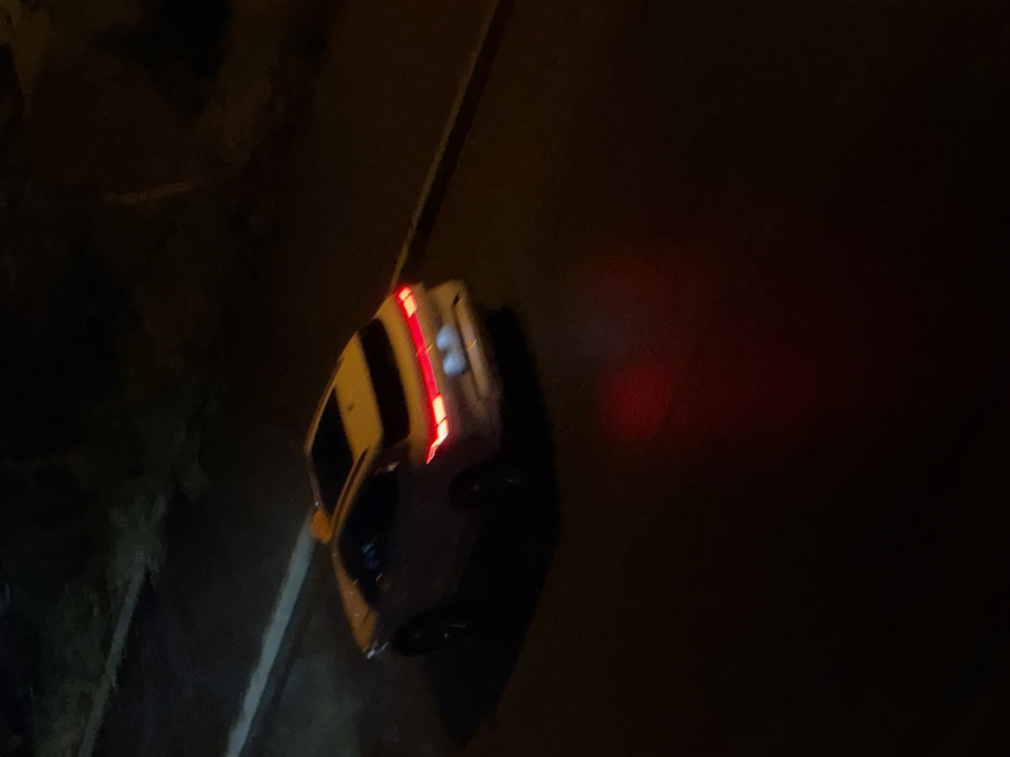 At night, a white SUV with its brake lights illuminated is seen from a high angle on a dark, paved road. The vehicle has a sunroof and a yellow object on its roof, suggesting it may be a taxi or ride-sharing vehicle. The road is bordered by a curb and a dimly lit area with vegetation. A red reflection is visible on the road surface behind the vehicle. The setting appears to be an urban environment, possibly Tbilisi, Georgia, given the visual context.