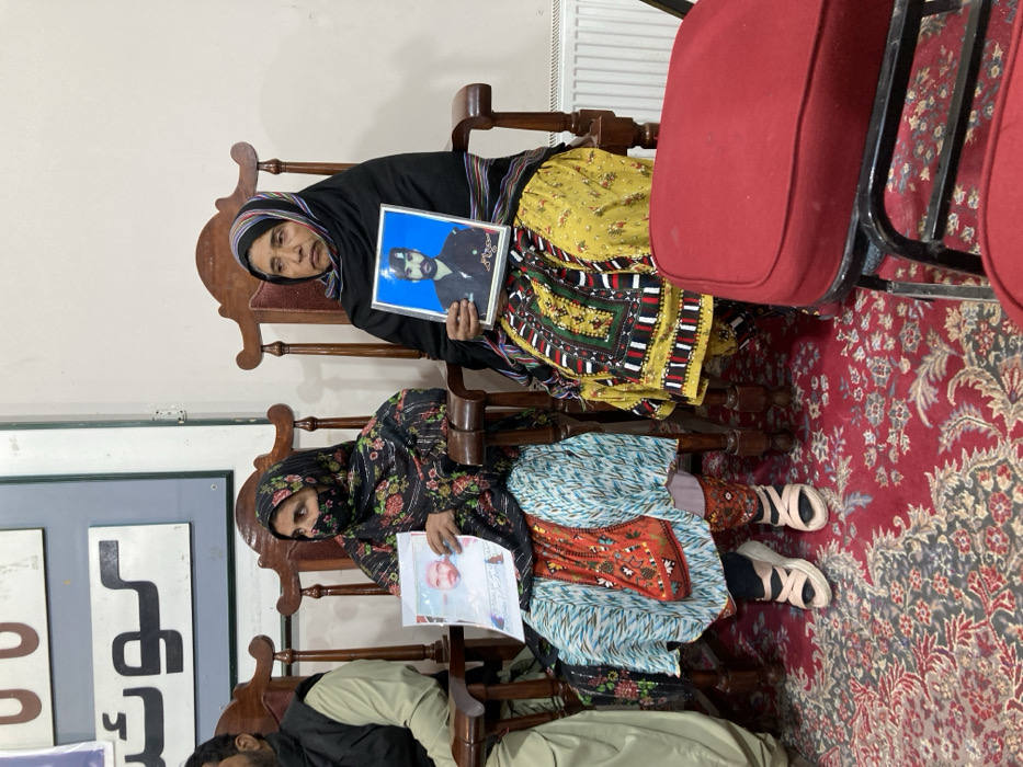 An indoor scene in what appears to be a public or official building in Quetta City Tehsil, Pakistan, based on visible Urdu script "کوئٹہ" (Quetta) on a background sign. Two women are prominently seated in ornate wooden chairs on a red patterned carpet. The woman on the left, wearing a black floral head covering and a patterned dress, holds a paper displaying a portrait of a clean-shaven man. The woman on the right, wearing a black shawl and a yellow embroidered dress, holds a framed portrait of a bearded man in a military-style uniform. Both women exhibit solemn expressions. A man's torso is partially visible on the far left. The background consists of a light-colored wall with a mounted sign and a white radiator. A red-cushioned chair with a black metal frame is in the foreground on the right.