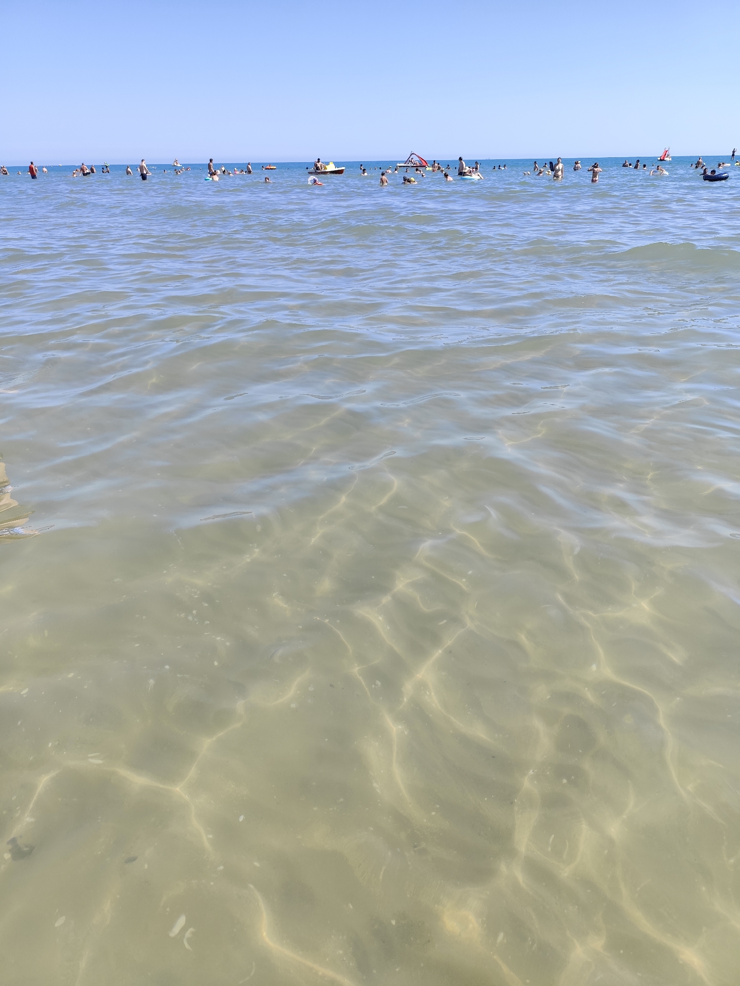 The image depicts a crowded beach scene in Rimini, Italy, on a clear, sunny day. The foreground is dominated by shallow, clear ocean water, revealing a sandy bottom with visible undulations and light patterns caused by the sun's reflection. In the midground and background, a multitude of people are enjoying the water. Some are swimming or wading, while others are floating on various inflatable devices, including tubes and a small boat. Several inflatable slides are also visible in the water. The horizon line is clearly defined by the meeting of the blue sky and the distant sea. The overall atmosphere is one of leisure and recreation.