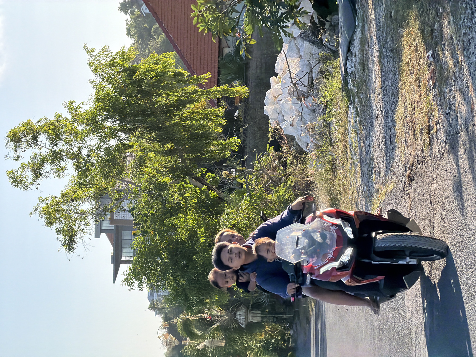 The image shows a family of four riding a red and black scooter on a paved road in Kuta Selatan, Indonesia. A man is driving the scooter with a young child seated in front of him. Behind the driver, a woman is seated, holding another young child on her lap. The family is dressed in casual clothing. The background features lush green trees and foliage, a modern building with large windows, and traditional Balinese architecture with red tile roofs. There is also a pile of white rocks or building material on the right side of the road. The scene is illuminated by sunlight, casting shadows on the road, suggesting it is either early morning or late afternoon.