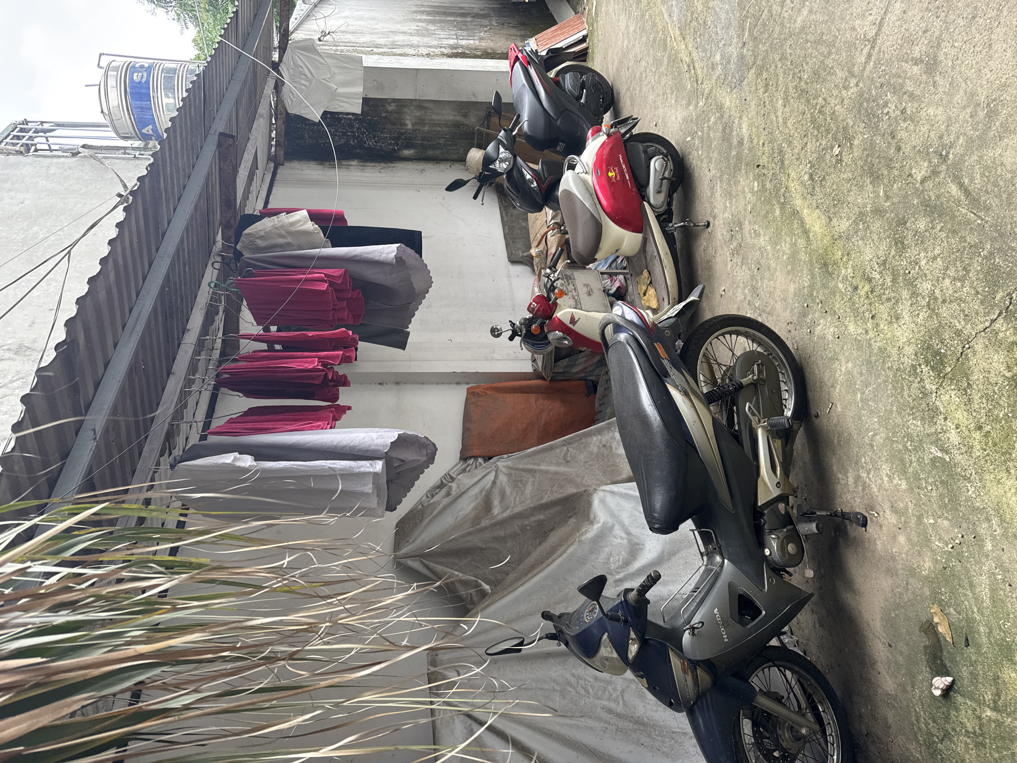 The image shows an outdoor alleyway in Hà Nội, Vietnam. Several motorbikes are parked along the right side of the alley. Clothes are hanging on a line to dry on the left. A water tank sits on the roof of the building. The ground is concrete with patches of moss.