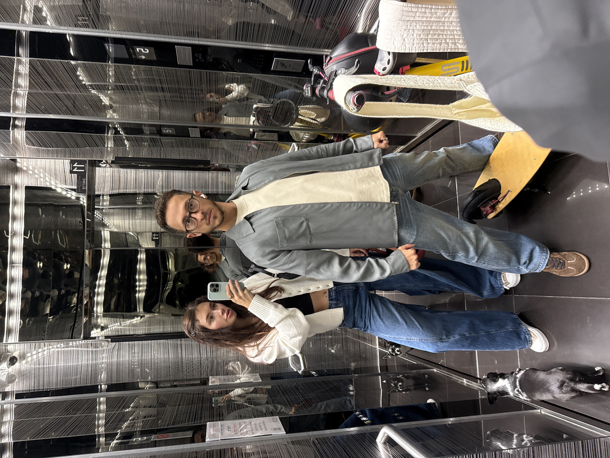 A couple, a man and a woman, are standing in what appears to be a modern elevator with reflective walls. The woman is taking a photo of herself and the man using her smartphone. The man is wearing glasses, a light-colored shirt, a grey jacket, and blue jeans. The woman is wearing a white cardigan over a dark top, blue jeans, and sneakers. There is a small black and white dog lying on the elevator floor. An unseen object, is also present within the elevator space with them. The elevator walls have a unique design featuring vertical strands. It is not possible to determine the location.