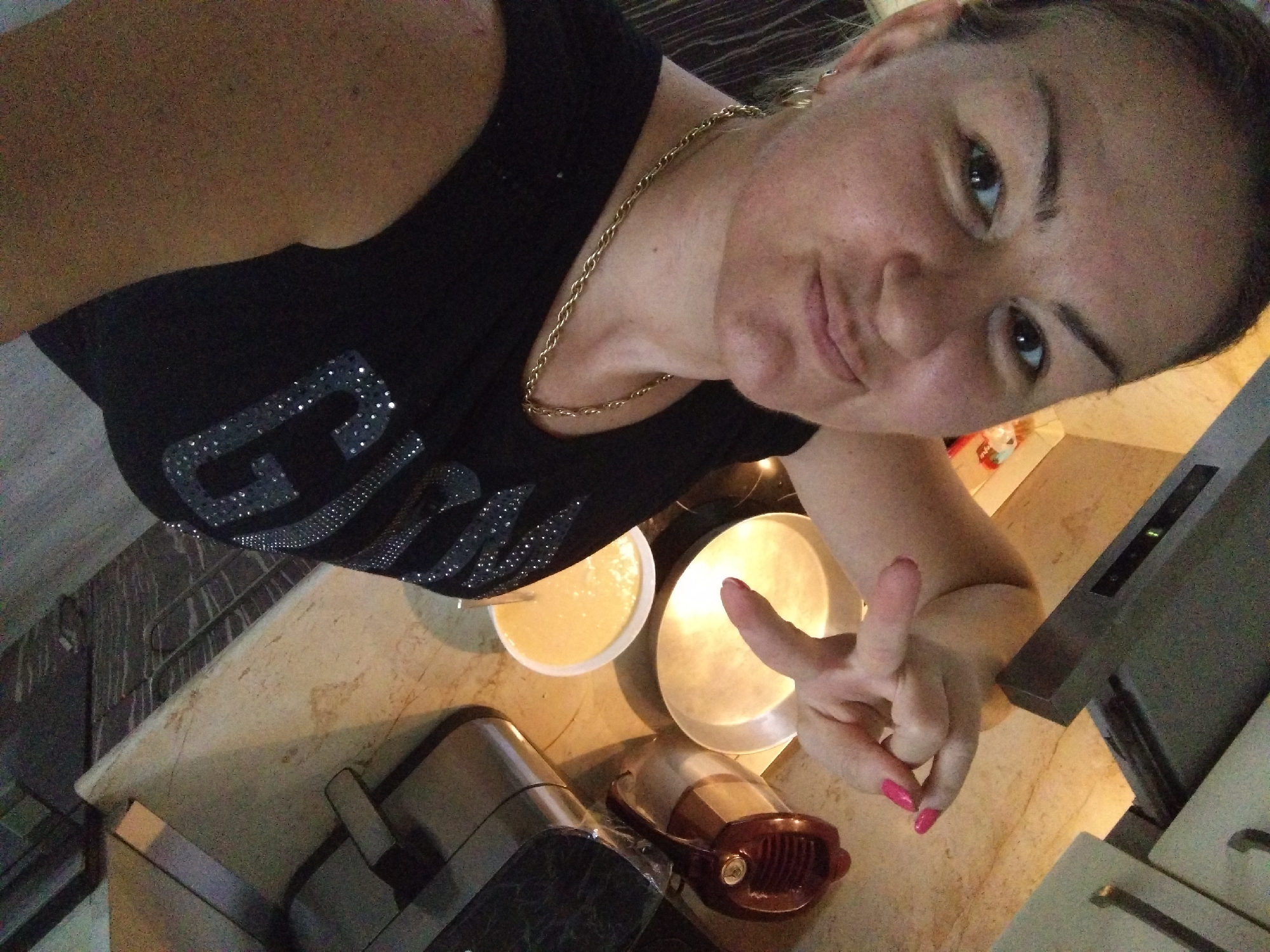A selfie photograph taken indoors, likely in a kitchen. The subject is a woman with fair skin, wearing a black shirt with sparkly decorations and a gold chain necklace. She is positioned at an angle relative to the camera, looking directly at it with a slight smile. Her left hand is visible near a countertop, gesturing with the thumb and index finger extended. On the countertop are several items, including a white bowl containing a light-colored liquid, a round metal plate, a small brown-colored appliance, and a dark-colored appliance with a transparent wrapping. In the background, part of a kitchen cabinet is visible. The location is not explicitly revealed by the image, so it would be impossible to establish definitively that the image was taken in Stara Zagora, Bulgaria.