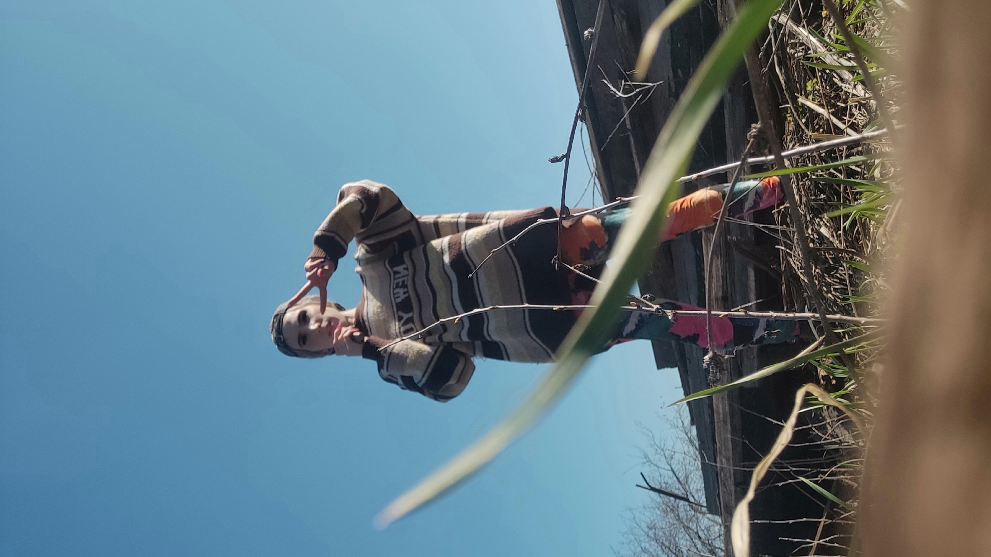 A low-angle shot depicts a person wearing a horizontally striped sweater with "NEW YORK" vertically printed on the front, making a peace sign with their right hand. They are wearing a hat and colorful pants. Above and to the left of the person is a clear blue sky. A wooden structure with foliage is visible to the right, including what appears to be laundry hanging on a line. Based on the context clues, the location is likely Chaikovsky, Russia.