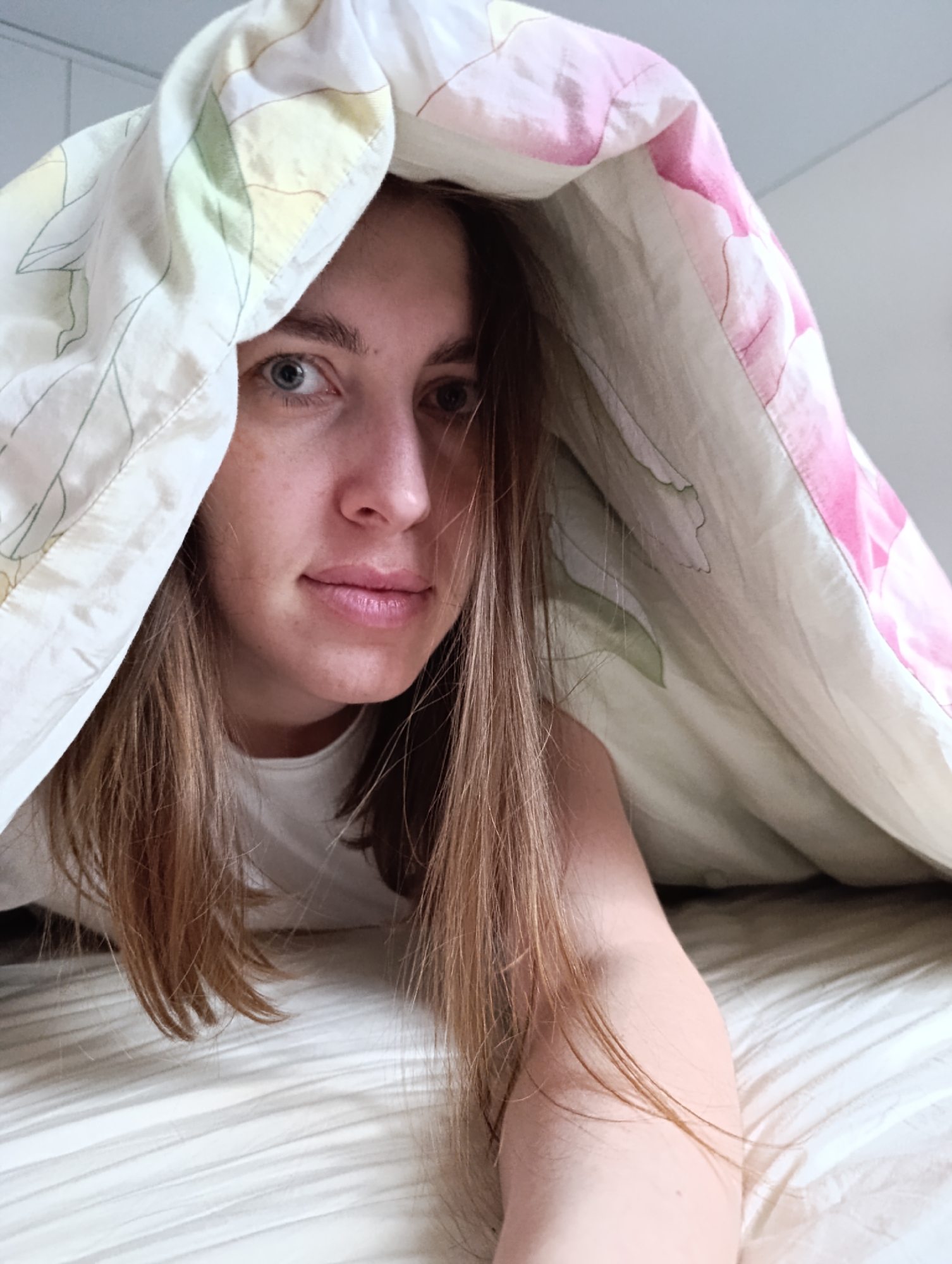 A woman is lying in a bed, covered by a floral-patterned duvet. The woman has light brown hair and blue eyes. The location is unknown.