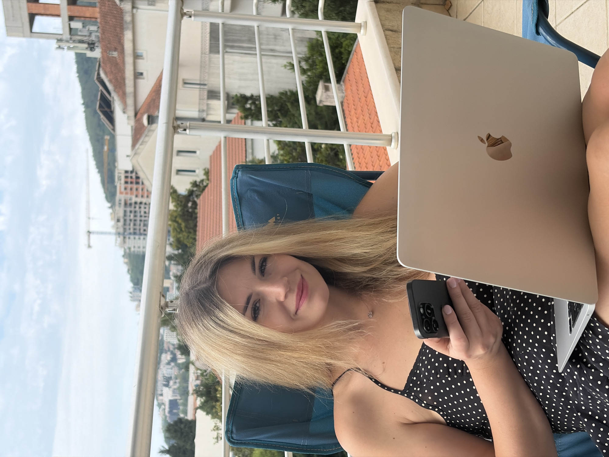 A young woman with long, blonde hair is seated in a blue outdoor chair on a balcony, looking directly at the viewer with a subtle smile. She is wearing a black dress adorned with white polka dots and delicate spaghetti straps, along with a small pendant necklace. In her left hand, she holds a dark-cased smartphone with a visible multi-lens camera array, while a silver Apple MacBook rests on her lap or beside her, displaying its iconic Apple logo.
The setting is a balcony, framed by white metal railings in the foreground. Beyond the railing, a picturesque view unfolds, characteristic of Budva, Montenegro. Numerous buildings with distinct red tiled roofs are visible amidst lush green trees. In the mid-distance, a city landscape extends, with hills covered in vegetation rising in the background. A tall construction crane stands out against the bright, light blue sky, which features a few scattered white clouds. The scene suggests a relaxed daytime activity, possibly working or browsing, in an attractive outdoor environment.