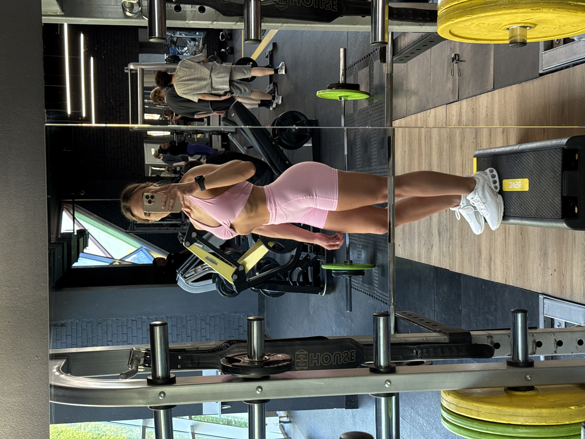A young woman, with blonde hair and wearing a pink athletic set, stands in a gym and takes a mirror selfie. She is positioned in the foreground, with a barbell rack to her left and a portion of another barbell rack to her right. In the background, visible in the mirror's reflection, other gym-goers are present, with some engaging in weightlifting activities. The gym environment features various weightlifting equipment, including barbells, weight plates (some green and yellow are visible), and weight machines. The gym's walls are a mixture of dark brick and lighter colored surfaces, with some decorative elements in the ceiling area. The floor appears to be a combination of rubber matting and wooden platforms. This scene is located in a gym in Saint Petersburg, Russia.