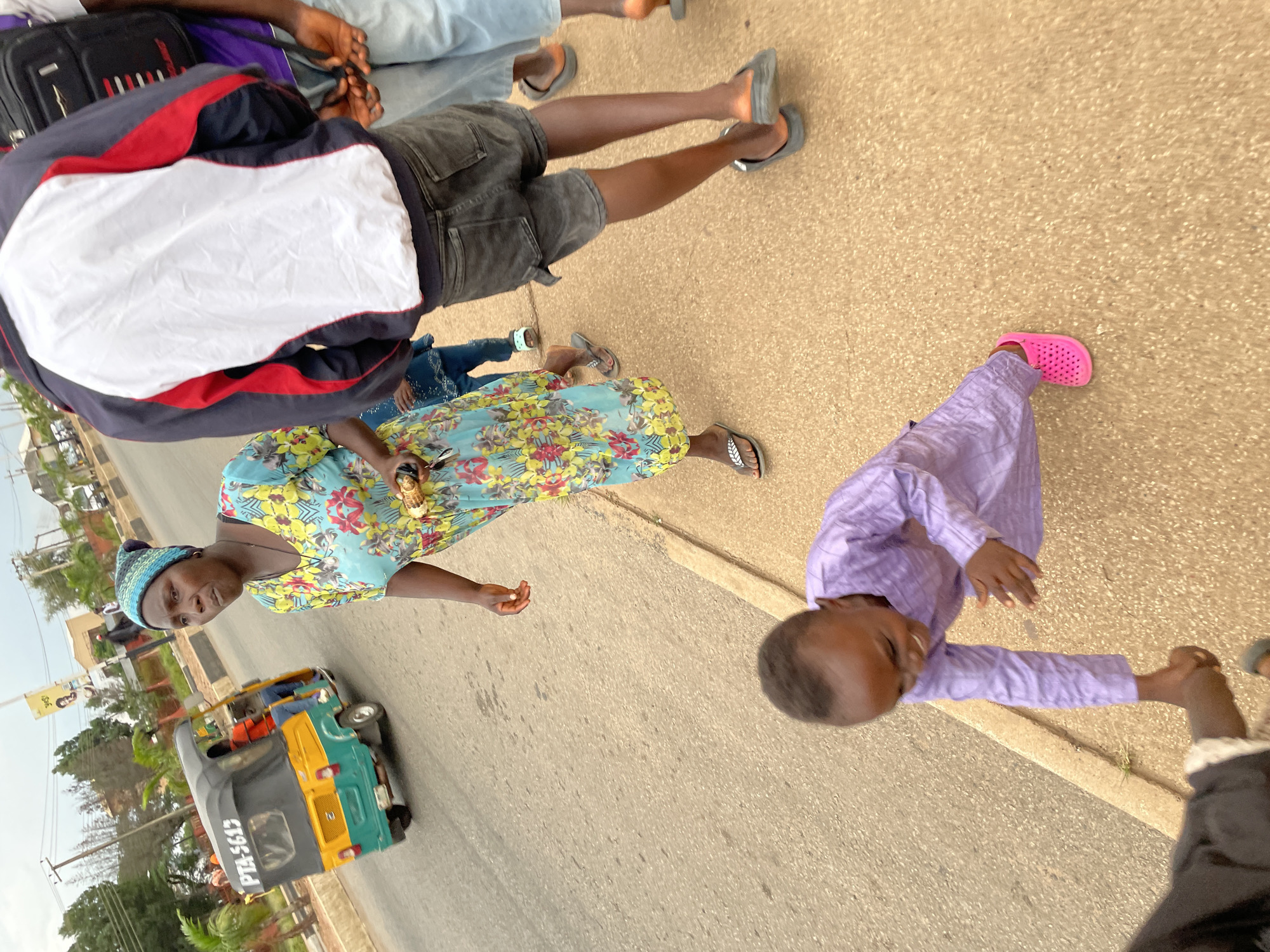 In Jos, Nigeria, a woman in a colorful floral dress and a blue and green headscarf walks on a sidewalk adjacent to a street. She carries a small object in her hand. To her left, a small, green and yellow auto-rickshaw, labeled "PTA-5613", is driving away. A young child in a purple outfit is on the ground in front of her, reaching out to someone on the far left, who is only partially visible. Other people, including a person in a jacket and shorts, and a person in a purple shirt and jeans, are walking on the sidewalk behind the woman. The environment appears to be an urban setting with buildings and trees visible in the background. The ground is paved with asphalt and concrete.