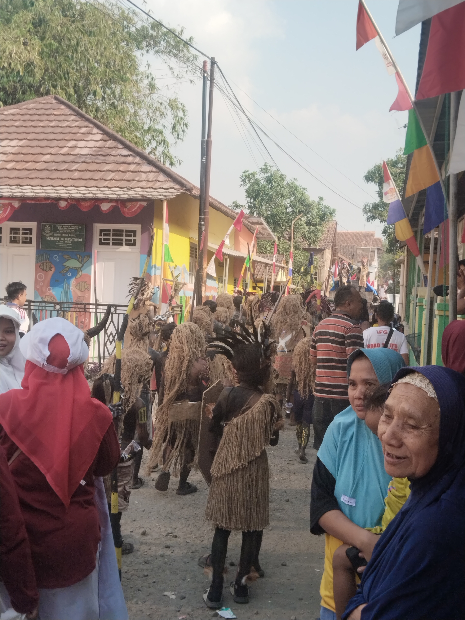 The image depicts a lively street scene in Jembayat, Indonesia, likely during a cultural event or celebration. A procession of individuals, many adorned in traditional costumes resembling grass skirts and headdresses, are walking down a dusty street lined with buildings. The buildings are painted in bright colors and decorated with flags, indicating a festive atmosphere.
In the foreground, several people are visible, including a woman in a blue hijab, an elderly woman with a warm smile, and children. Their presence suggests they are either participants or spectators of the event. The crowd appears dense, with people of various ages and backgrounds interacting and observing the parade.
The environment is characterized by a clear, bright sky, suggesting it's daytime. Overhead wires and electrical poles are present, typical of urban or semi-urban settings. The ground is unpaved, a common feature in many parts of Indonesia. The overall impression is one of a vibrant community gathering, filled with cultural expression and social interaction.
