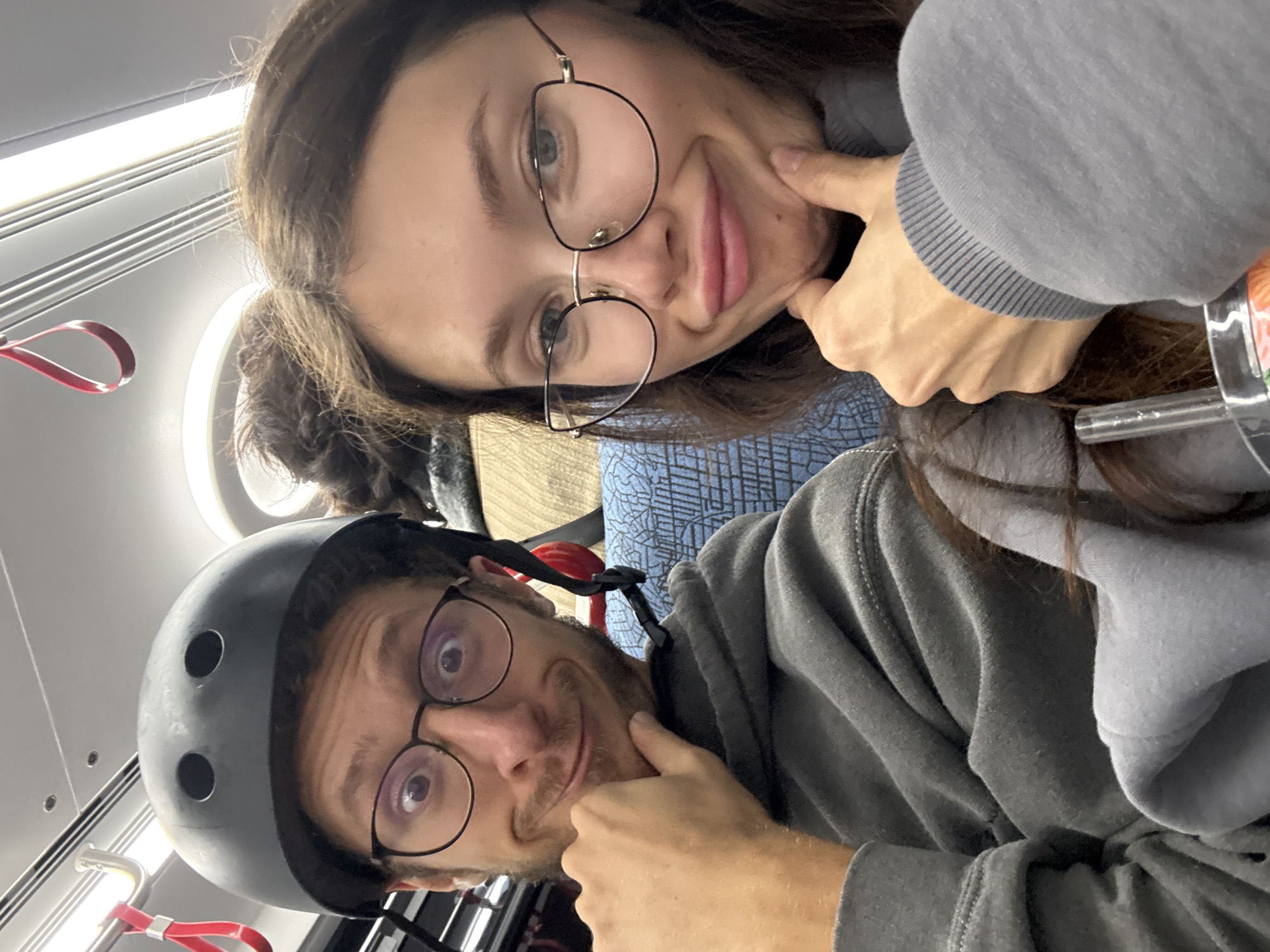 A rotated selfie captures two young adults, a man and a woman, making playful expressions. Both are looking towards the camera, each pressing a finger into their cheek to create a squished-face effect, conveying a cheerful and lighthearted mood.
The woman, positioned in the upper part of the frame, has long, dark hair and light-colored eyes. She wears large, round, dark-framed glasses and is dressed in a grey hoodie over a blue top with a subtle geometric pattern.
The man, in the lower part of the frame, has short facial hair, round glasses, and is wearing a dark grey helmet with ventilation holes. He is also dressed in a grey hoodie.
The setting appears to be the interior of a public transport vehicle, likely a train or light rail, consistent with the Tel Aviv, Israel context. The background features bright, linear fluorescent lights on the ceiling, along with red hanging straps for standing passengers and a metallic overhead structure. No text is visible in the image.