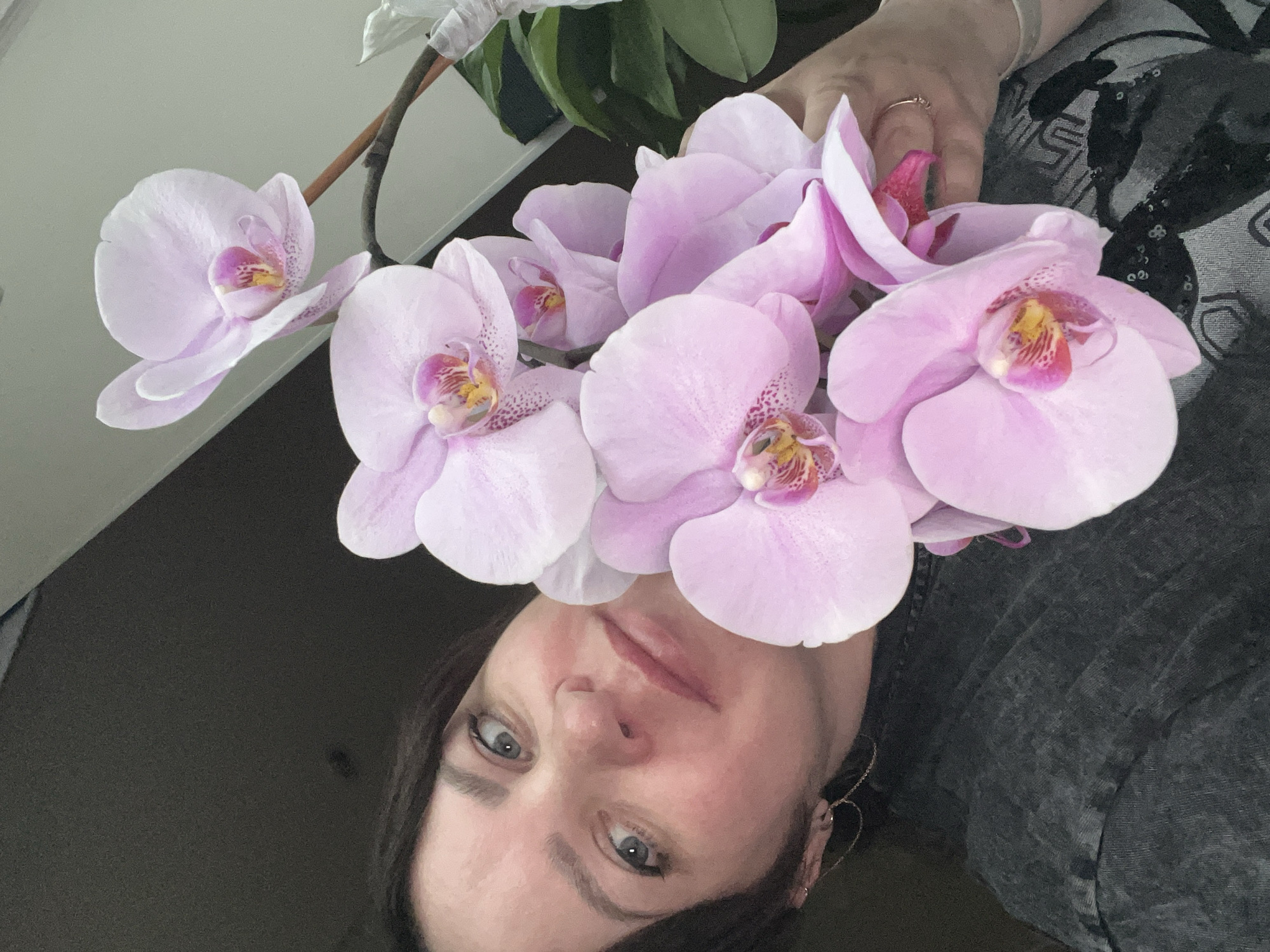A person with fair skin, dark hair, and light eyes holds a bouquet of light pink orchids up to their face. They wear a dark gray top with some sequins. Part of another orchid plant is visible in the background. No information about the location in Perm, Russia, can be discerned from the image.