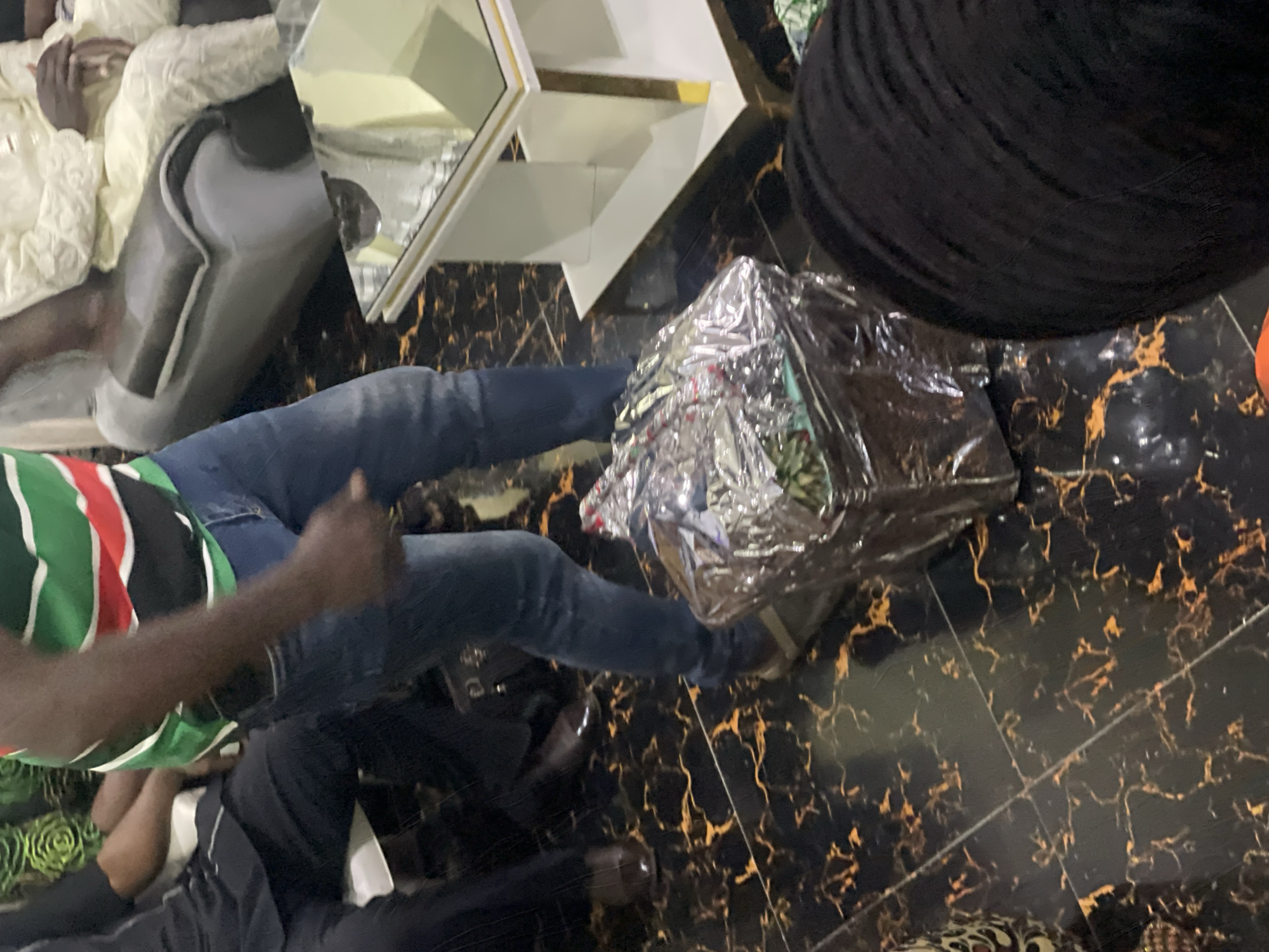 The image depicts an indoor scene, possibly a living room, with people seated on furniture. The floor is tiled with a black and gold marble pattern. In the foreground, a person wearing blue jeans and a green, red, and white striped shirt is partially visible, standing next to a silver-wrapped gift. Another person, wearing a black garment, is seated on the left. In the background, individuals are seated on a sofa. A modern coffee table with a glass top and white and gold accents is positioned near the sofa. The overall lighting suggests an indoor setting, likely during the daytime. The presence of a wrapped gift suggests a celebratory occasion.