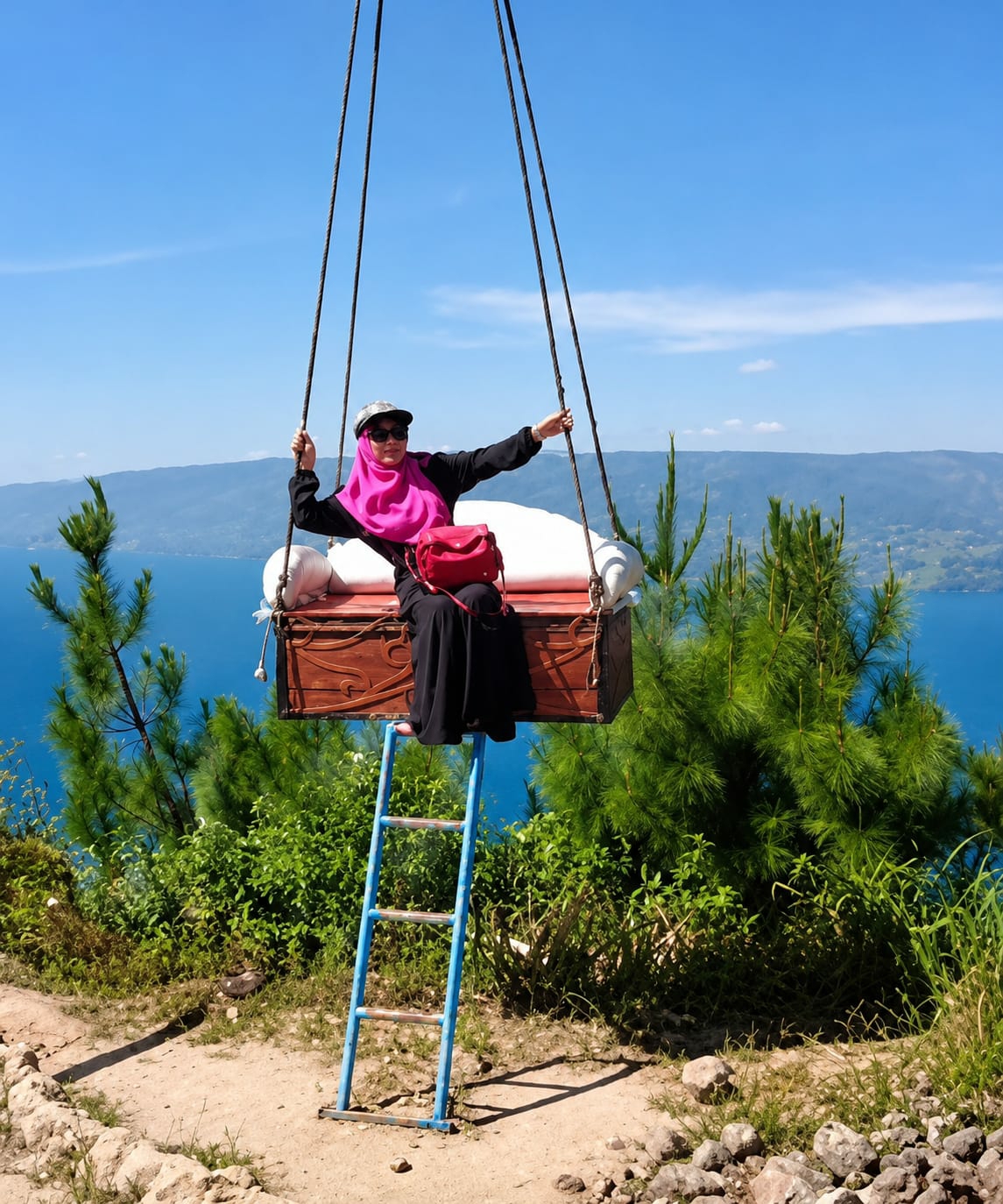 A person wearing a pink hijab, a black abaya, and sunglasses sits on a wooden chest suspended by ropes. The person is looking to their right with their arms outstretched. A red bag is slung over their left shoulder. A blue ladder is positioned below the chest, leaning against it. The chest is situated on a hillside with green vegetation and trees. In the background, a large body of water is visible, with hills or mountains beyond it under a clear blue sky. The scene appears to be an outdoor attraction, possibly for photography, in Parik Sabungan, Indonesia.