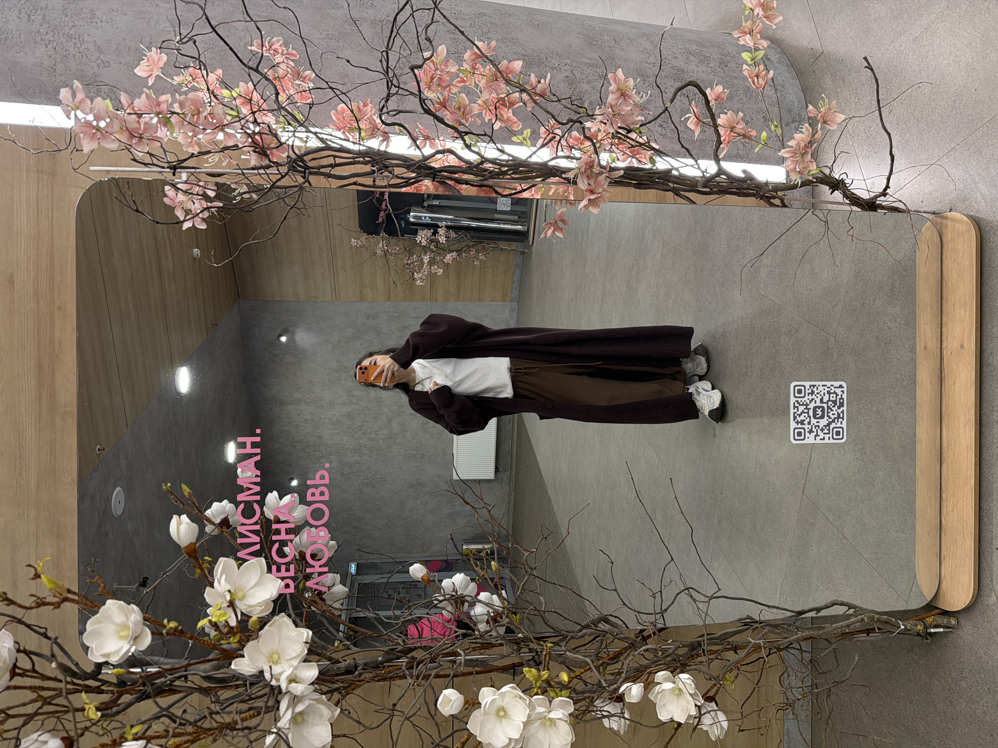 An indoor scene captures a person taking a mirror selfie, with the image rotated 90 degrees counter-clockwise so the person appears horizontal. The central focus is a large, rectangular mirror, partially framed by intricate artificial branches bearing both delicate pink blossoms and larger white magnolia-like flowers.
The person, reflected in the mirror, is wearing a long, dark brown coat or cardigan over a white top, brown pants, and white sneakers. They are holding an orange-cased smartphone in front of their face to capture the reflection.
The background visible within the mirror's reflection reveals a modern interior with smooth grey walls and some sections of light wooden paneling. Prominently displayed on one of the grey walls, reflected in the mirror, is bright pink neon text that reads "ТАЛИСМАН. ВЕСНА. ЛЮБОВЬ." (which translates to "TALISMAN. SPRING. LOVE."). To the right of the person's reflection, a black and white QR code is affixed to the grey wall. The lighting appears to be artificial, coming from ceiling fixtures, and the overall setting suggests a chic commercial or public space designed with decorative flair. The time of day is not discernible.