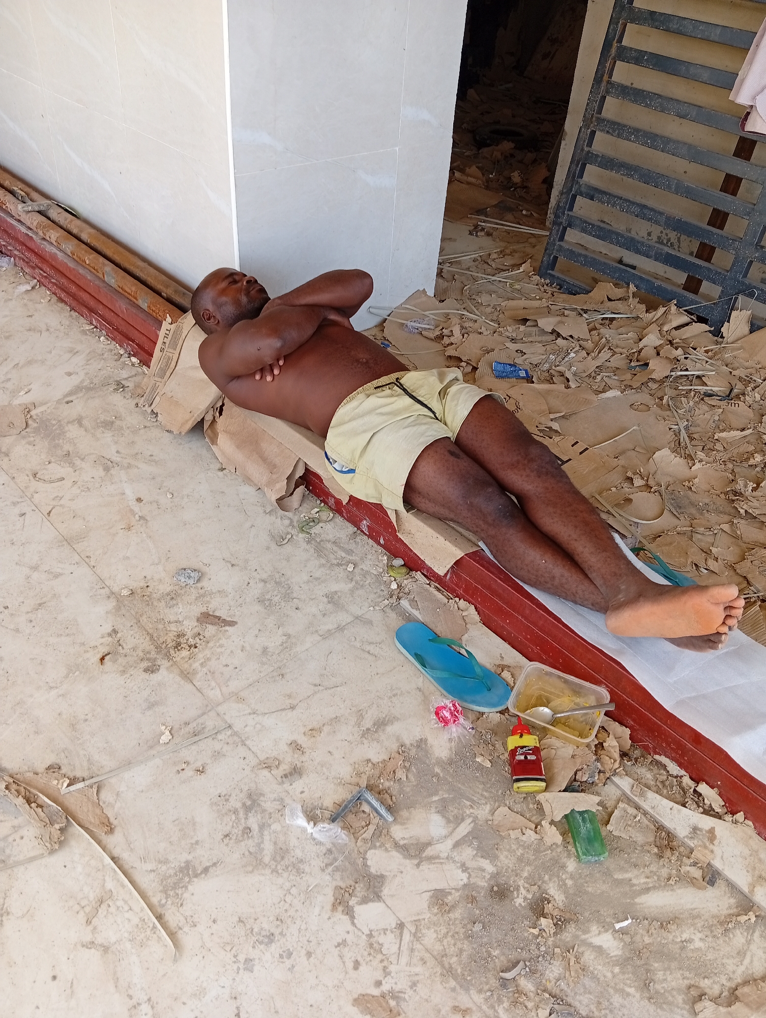 A shirtless, dark-skinned man wearing light-colored shorts is lying on his back with his arms crossed on a flattened cardboard box on a tiled floor. He is positioned alongside a red-painted metal beam next to a stack of pipes leaned against a wall in what appears to be a construction or renovation site. Around him are scattered debris, including cardboard fragments, packaging materials, and personal items like a pair of blue flip-flops, a clear plastic container holding food with a spoon, a small bottle with a red label, and a green object. The location appears to be indoors, judging by the adjacent white wall and the presence of an open doorway leading to a darker area. Through the doorway is visible a metal security door and more construction debris. The setting appears to be a construction zone in Abuja, Nigeria.