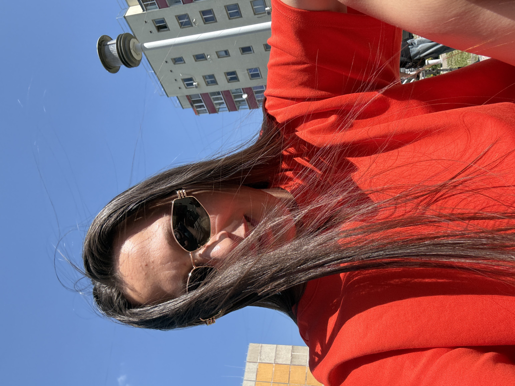 The image is an upward-angled shot of a woman wearing sunglasses and an orange top. She is outside, in what appears to be an urban setting. In the background, a tall building with a grid of windows is visible, along with a utility pole. The image was taken in Chaikovsky, Russia.