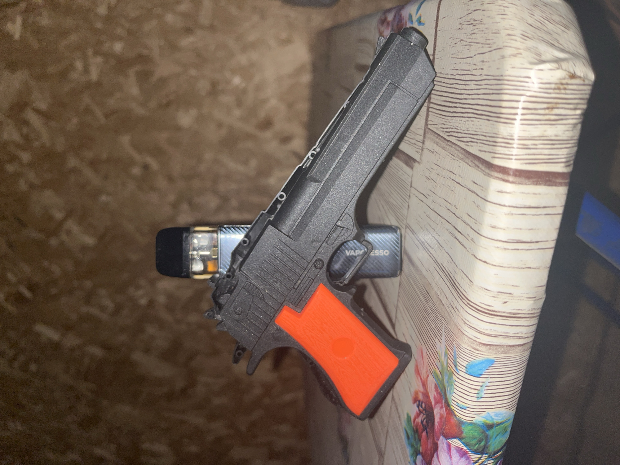 The image depicts a toy gun and a vape device resting on a table. The table has a wood-grain pattern and is partially covered with a floral-themed tablecloth. The background appears to be a textured, brown wall. There are no people or specific landmarks of Chaikovsky, Russia visible in the image.