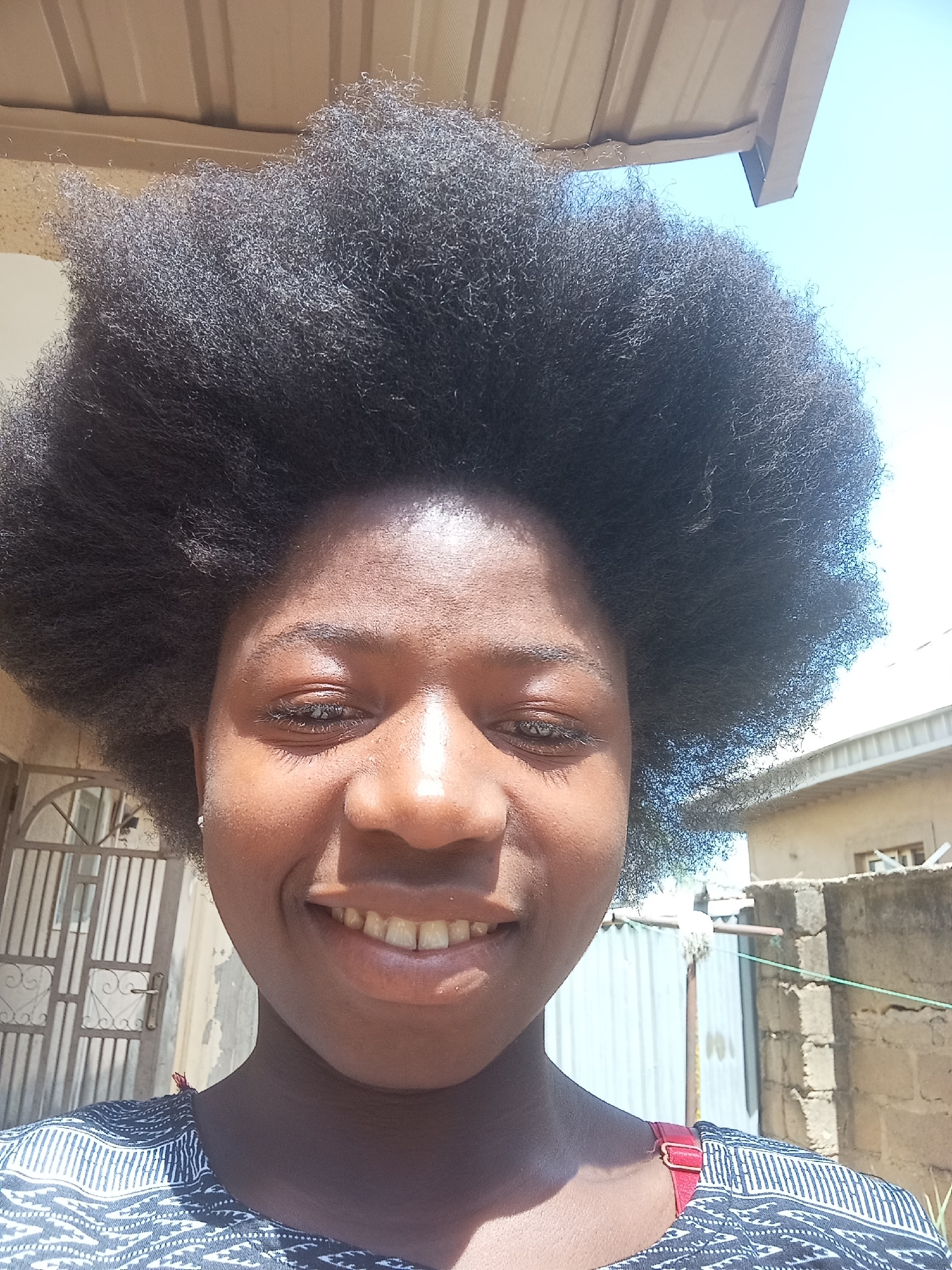 A smiling young Black woman stands outdoors in Jos, Nigeria. Her large afro is the dominant feature. She wears a patterned top. Behind her, a building with a metal roof and a partially visible gate can be seen. A second, smaller building with a galvanized fence is also in the background. The sky is visible.