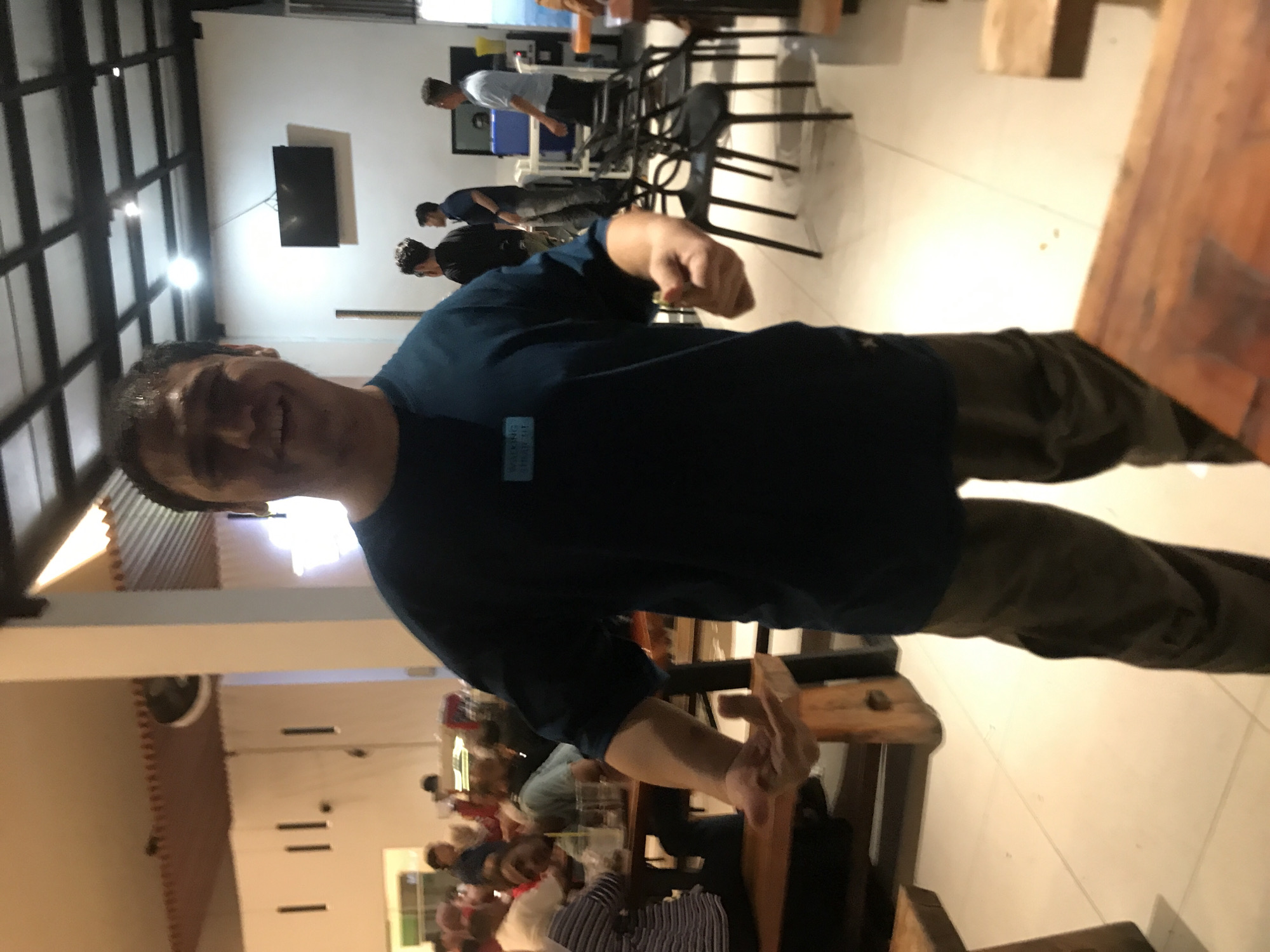 The image depicts a man in a dark blue shirt and brown pants, smiling at the camera while standing in what appears to be a bustling indoor restaurant or eatery. Other people are seen seated at tables behind him. The restaurant has a high ceiling with black metal beams. A flat-screen television is mounted on a wall in the background. The scene is likely in Lhokseumawe, Indonesia, based on the prompt's location specification. The furniture includes wooden tables and chairs. Patrons are engaging in dining and conversation. A ceiling fan is visible.