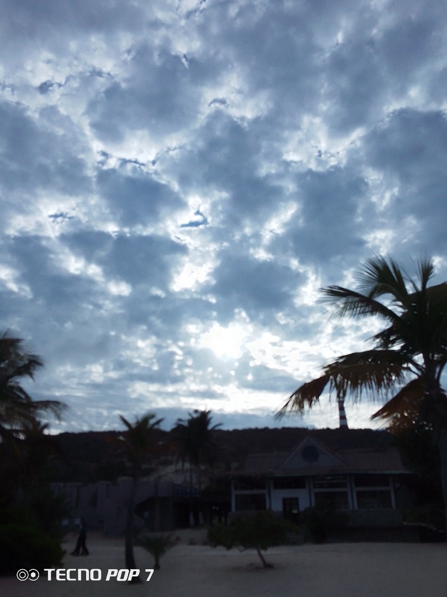 The image depicts a beach scene in Pedro González, Venezuela. A building is in the middle ground. Palm trees are scattered throughout the scene. Above, there's a sky filled with textured, gray clouds with sunlight filtering through. At the bottom left corner is a "TECNO POP 7" watermark.