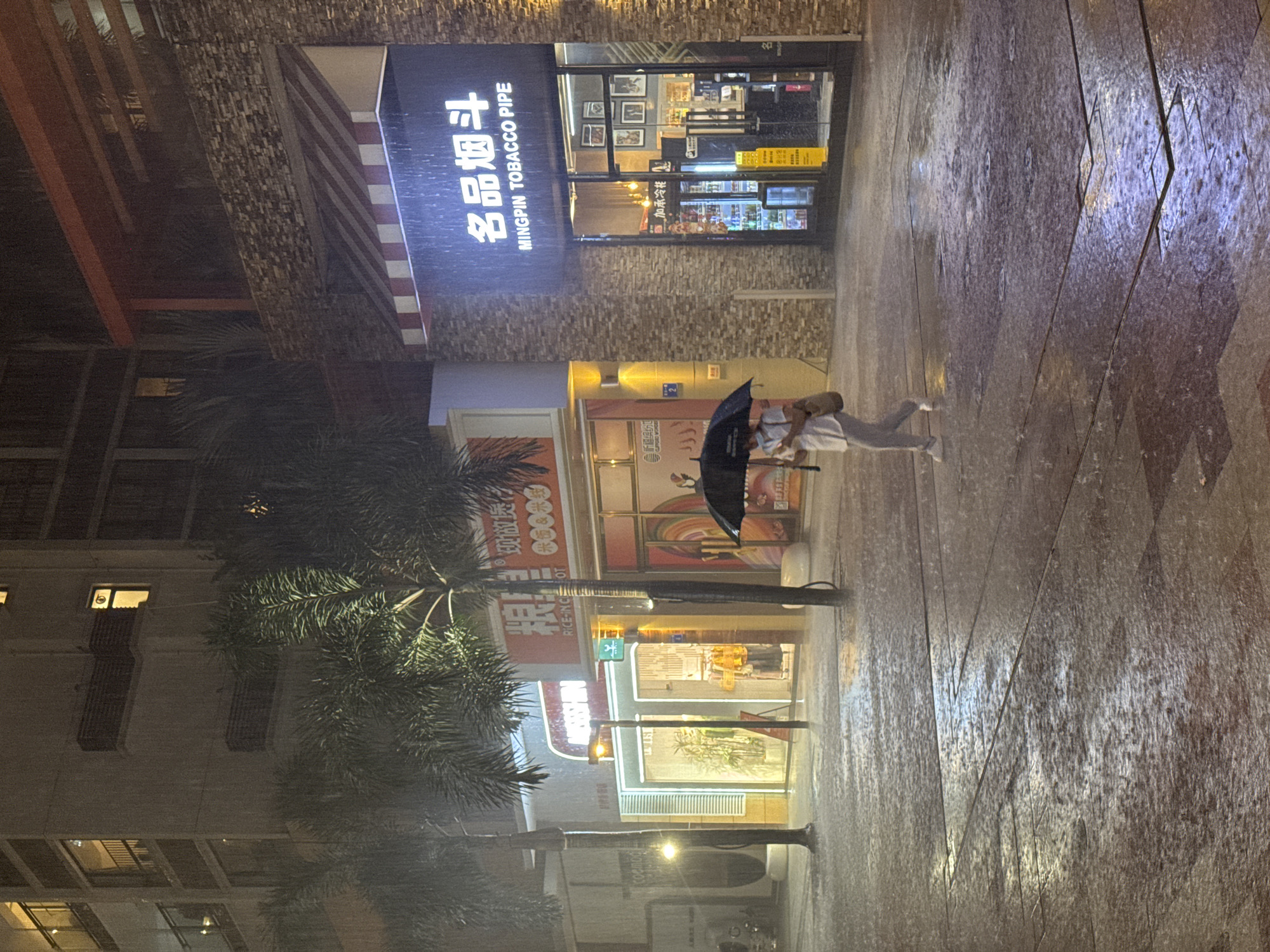 A street scene in Panyu District, China, during a heavy rain, features a pedestrian with an umbrella walking past several storefronts. The stores include a tobacco pipe shop with the sign "MINGPIN TOBACCO PIPE" and a restaurant called "RICE-INCE OT" that offers freshly cooked rice. A palm tree is visible on the left. The environment is wet, and the ground is reflective due to the rain. The image is shot at night, with artificial lighting illuminating the stores and streets.