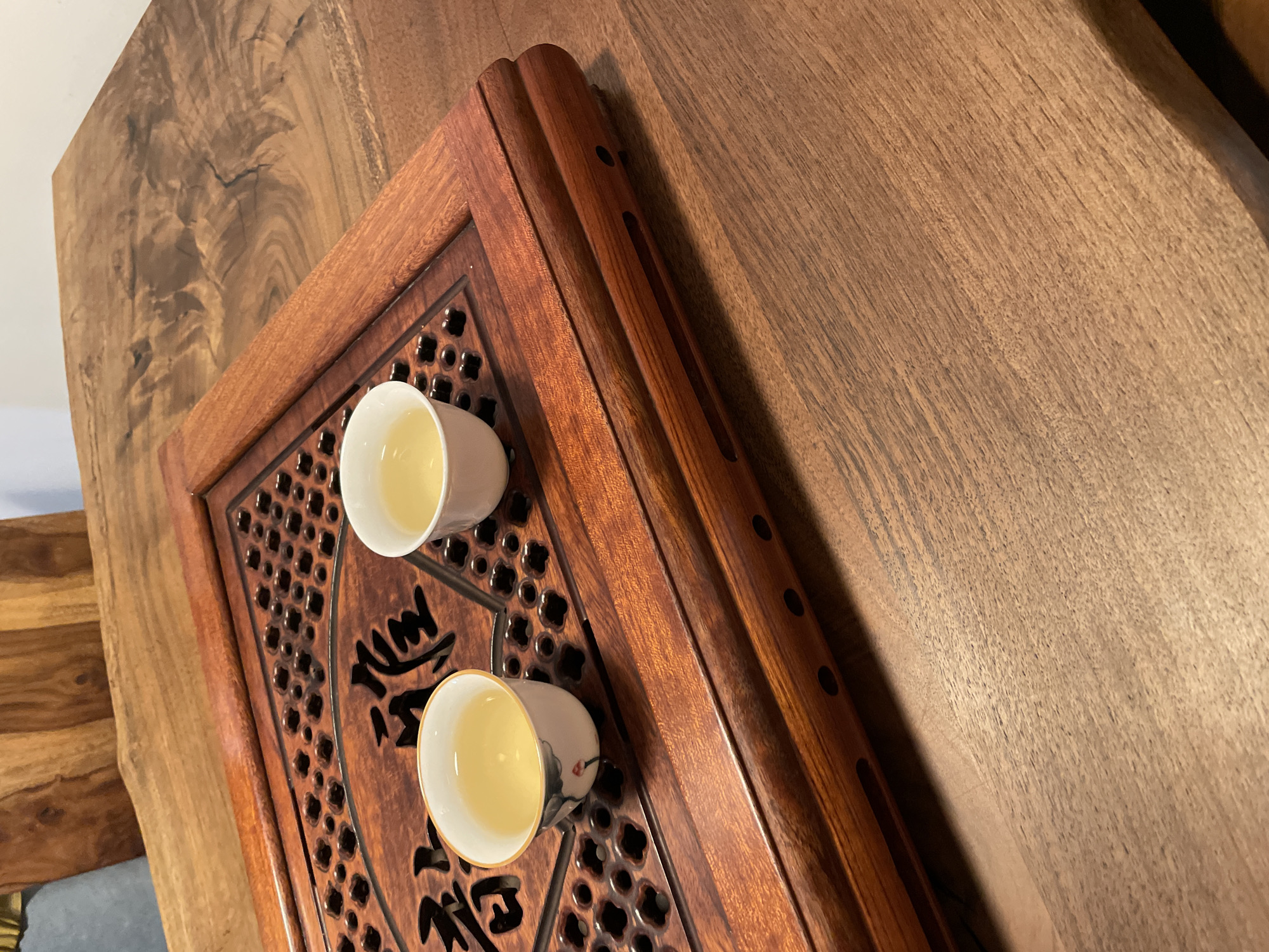 The image presents a close-up, angled view of two white porcelain teacups filled with a light yellow liquid, presumably tea. These cups are placed on a dark brown wooden tea tray featuring intricate carved designs, including a geometric lattice pattern and stylized Chinese characters, possibly calligraphy related to tea. The tea tray itself rests on a larger, lighter brown wooden surface, distinguished by its prominent, natural wood grain patterns, suggesting a substantial wooden table or counter.
The scene depicts an indoor setting, likely focused on the ritual of tea drinking or preparation. The soft, ambient lighting suggests it is daytime. While the image does not contain explicit location cues, it is set in Tbilisi, Georgia, as per the provided context. The arrangement evokes a sense of tranquility and an appreciation for traditional tea culture. No explicit emotional expressions are visible, but the setup conveys a calm atmosphere.