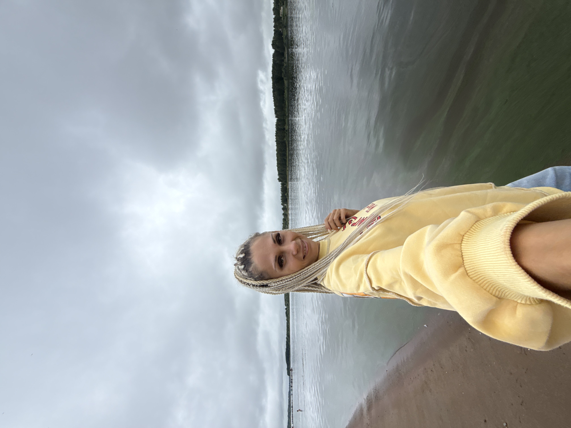A woman with blonde braided hair is taking a selfie on a beach next to a body of water. She is wearing a yellow sweatshirt. The sky is cloudy, and there is a line of trees in the distance across the water. The water appears murky, with some green algae.