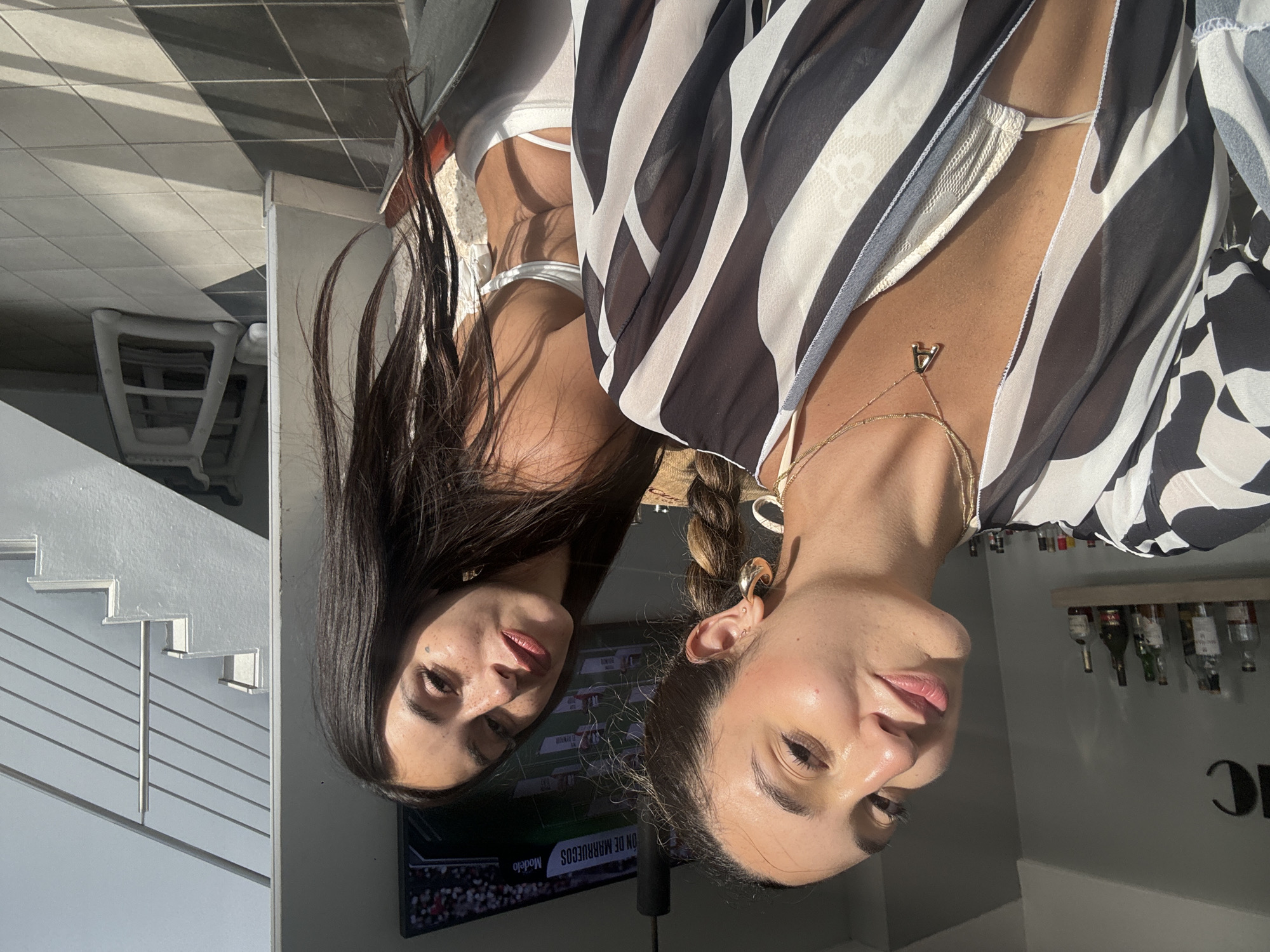The image shows two young women posing for a selfie. The woman on the left is closer to the camera, smiling with her head tilted. She is wearing a black and white striped sheer cover-up over a white bikini top and a gold necklace with an "A" pendant. The woman on the right is slightly behind and to the right, also looking towards the camera. She has long, dark hair and is wearing a white bikini top. Behind them, a television screen displays a sports broadcast. A shelf with bottles is visible on the left wall. To the right, a modern staircase with a silver railing leads upwards. The setting appears to be an indoor or semi-outdoor area with tiled flooring and natural sunlight casting shadows. The overall atmosphere suggests a casual, possibly vacation or leisure setting. The location is identified as San Juan, United States of America.