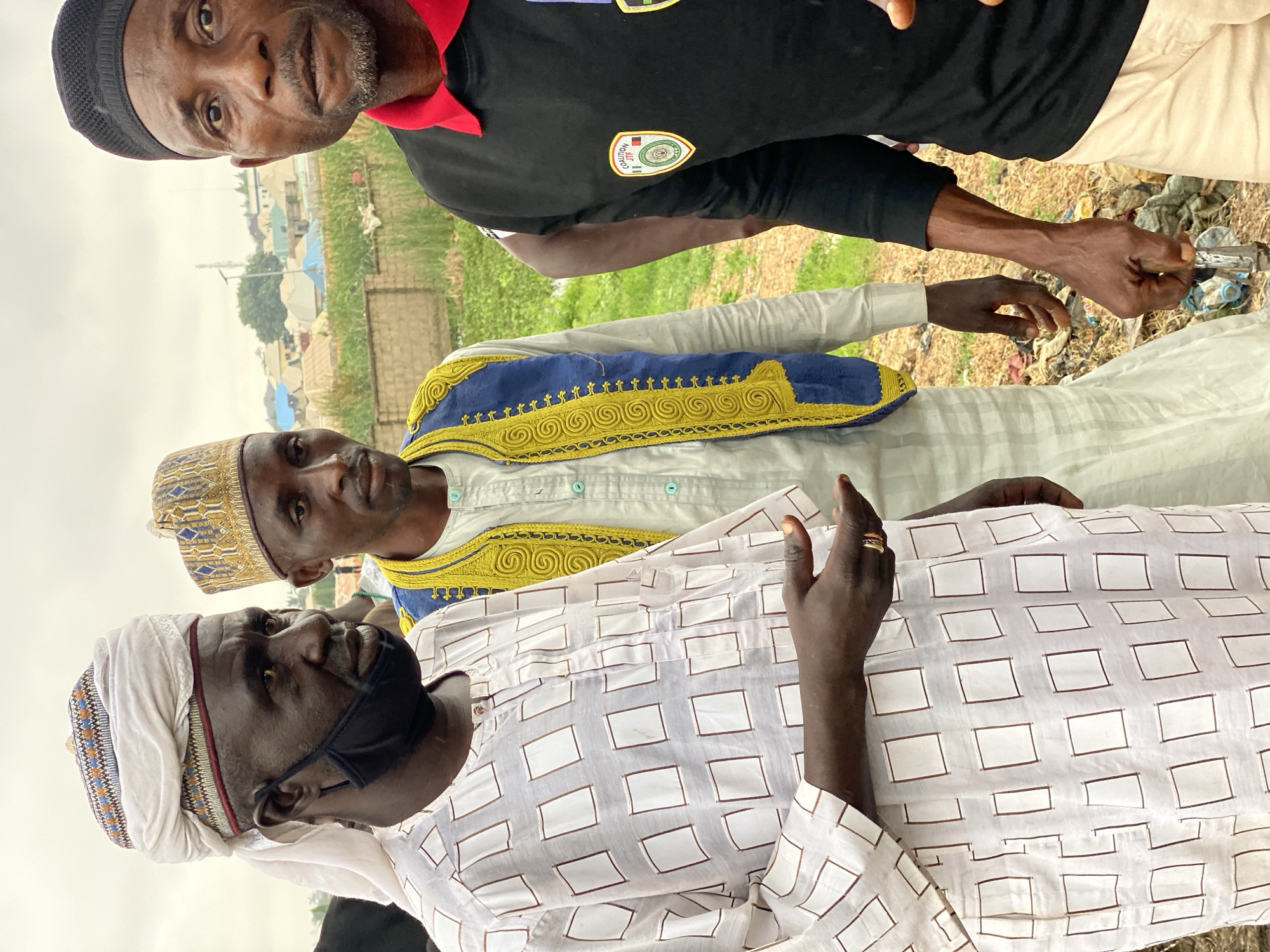 In the city of Kawo, Nigeria, three men are captured in a medium shot. The man on the left wears a white turban and a white tunic with a checkered pattern, and a black mask. His right hand is outstretched, showing a ring on his finger. The middle man sports a traditional yellow and blue embroidered vest over a white tunic and a gold-patterned cap. The man on the right, wearing a black t-shirt with a red collar and a black cap, stands close to the camera. His shirt features two circular patches. The background reveals a glimpse of a settlement with various structures, suggesting an outdoor urban or peri-urban setting. The ground is littered with debris. The sky is overcast.