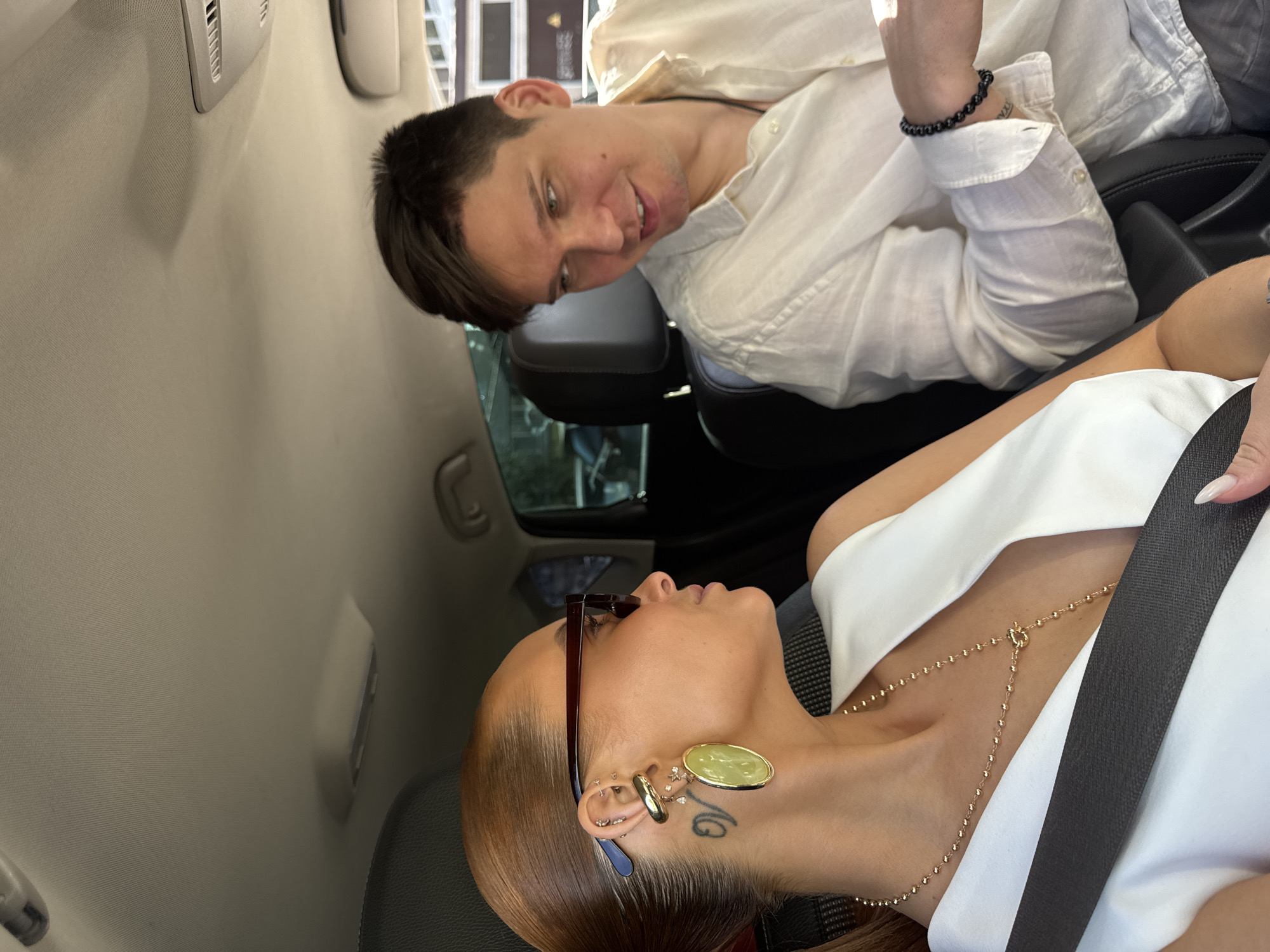Two individuals are inside a vehicle. A person in a white shirt is looking down at a person wearing a white top, sunglasses, a gold necklace, and a seatbelt. The person in the white top is looking up. The scene is likely inside a car, with the interior visible. The image does not provide enough context to determine if it is Chaikovsky, Russia.