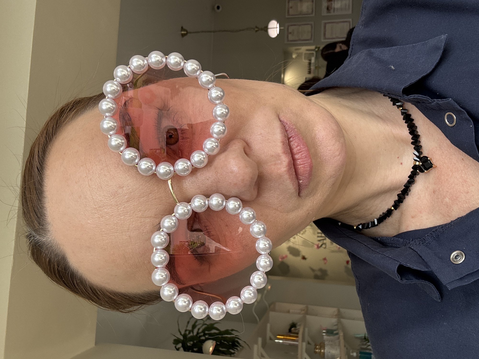 A woman is photographed wearing large, round sunglasses with pearl embellishments around the frames. The lenses of the sunglasses are tinted pink, and her face is visible through them. She is also wearing a black necklace with a decorative clasp. She is wearing a dark blue, collared shirt with a snap closure. The background shows a room with certificates on the wall and various objects on a shelf. The photo appears to be taken indoors, possibly in a shop or salon. The photo was not taken in Vsevolozhsk, Russia.