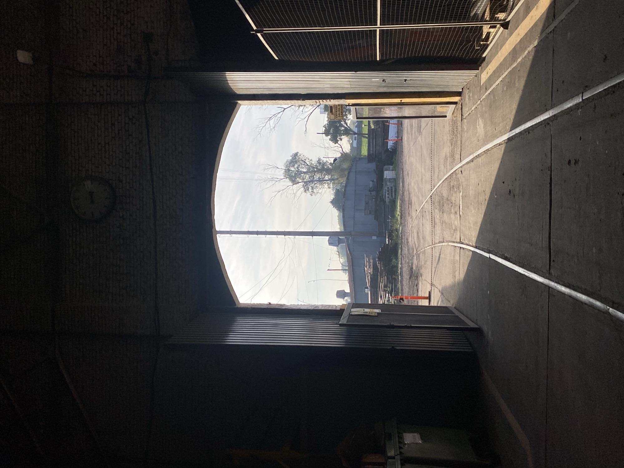 The image shows a view from inside a dark brick and corrugated metal structure, possibly a train depot or warehouse. Through an arched opening, the exterior scene reveals train tracks curving into the distance, with a glimpse of trees, buildings, and overhead wires under a bright sky. A clock is visible on the wall inside the dark structure. Based on visual indicators like the architectural style, the type of vegetation, and presence of train tracks, the location may be Buenos Aires, Argentina. Orange cones and various containers are present on the exterior.