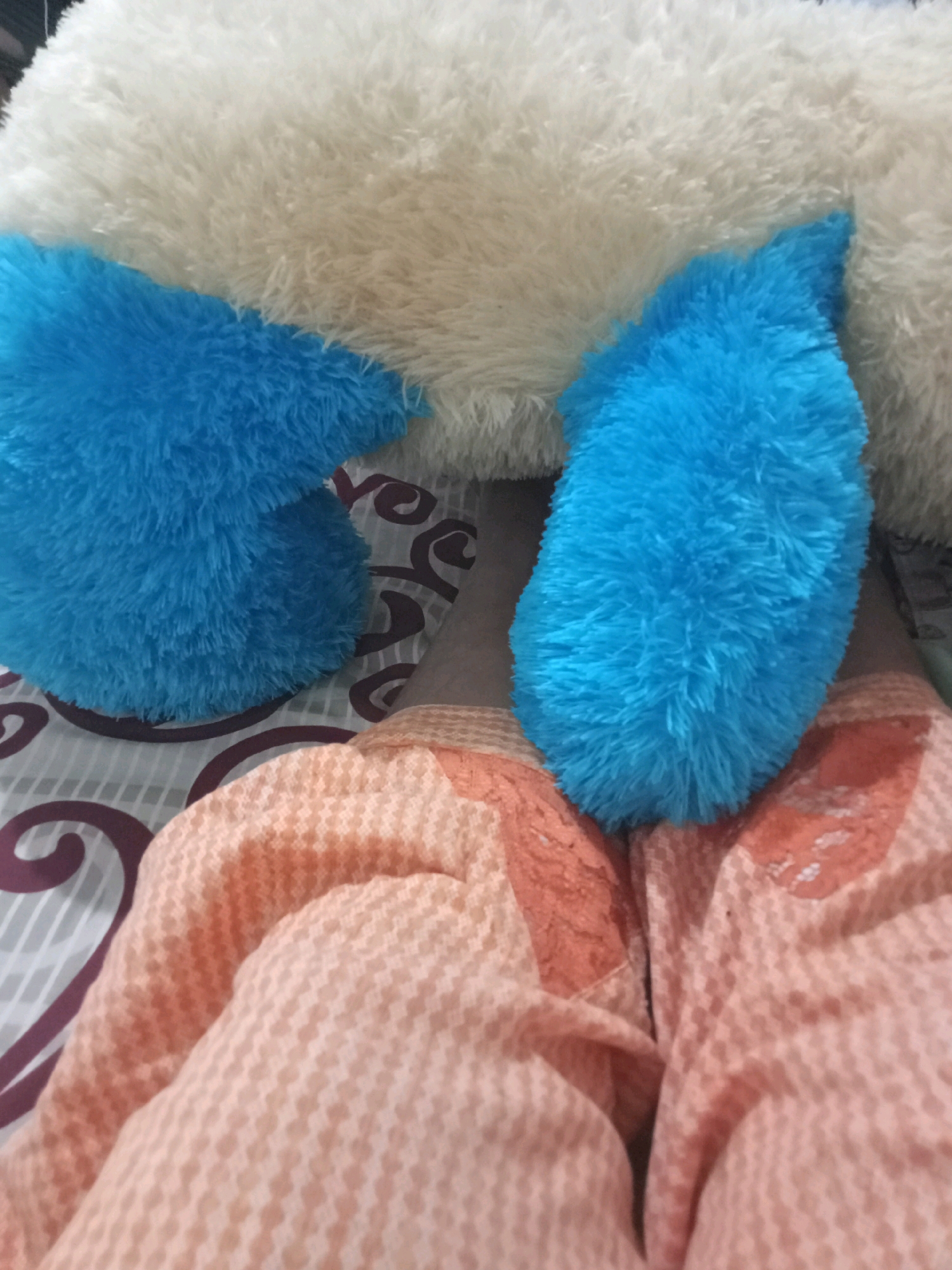 A close-up, overhead shot shows a person lying down, possibly in a bed, in Sukomananti, Indonesia. The person is covered by a peach-colored blanket with a small, woven pattern. Two large, fuzzy blue stuffed animal paws are resting on top of the blanket, partially obscuring the person's legs. Behind the paws is a large, fluffy white stuffed animal or pillow, which takes up the top portion of the image. The background bedding has a geometric pattern of gray lines and dark purple swirls.