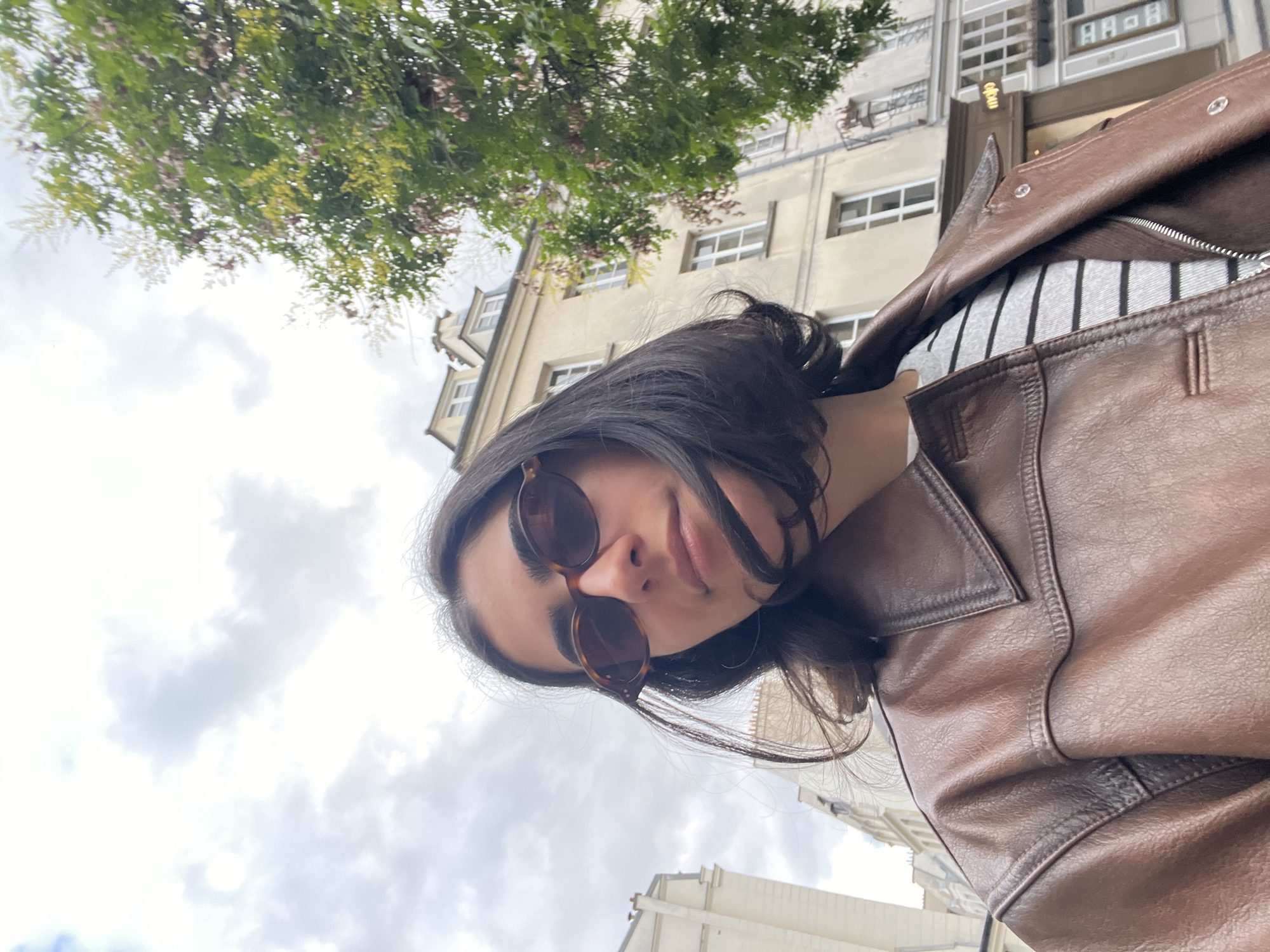 An individual is captured in a close-up, low-angle selfie, positioned outdoors against an urban backdrop on what appears to be an overcast day in Paris, France. The person, with dark, medium-length hair, is wearing dark brown, oval-framed sunglasses with a tortoiseshell pattern. They are dressed in a brown, textured leather-look jacket with a prominent collar and shoulder stitching, worn over a grey and white horizontally striped long-sleeved top.
The background consists of typical Parisian Haussmannian-style buildings with light-colored facades and numerous rectangular windows. On the left, a portion of a building with classical architectural details is visible. On the right, a multi-story building displays several white-framed windows and a dark, ornate lamppost or decorative fixture. A deciduous tree with green foliage and some reddish-brown or yellowish blossoms or new growth partially obscures the upper right section of this building. At street level on the right, a shopfront is visible, featuring a sign with the word "mojo" in a light-colored font above its entrance. The sky overhead is cloudy, dominated by white and light grey clouds. The individual's gaze is directed towards the camera, indicating a self-portrait.