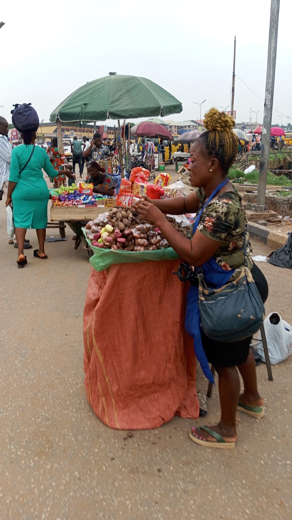 The image portrays a lively outdoor market scene in Onitsha, Nigeria, under an overcast sky, suggesting daytime hours. In the foreground, a female vendor with blonde braided hair styled in a high bun, wearing a floral patterned top, blue apron, and black shorts, stands behind her stall. She is actively arranging a prominent display of various goods, including a large mound of brown root vegetables or kolanuts, alongside smaller piles of light green fruits (possibly apples or garden eggs) and an assortment of packaged snacks and sweets in colorful, branded wrappers. The stall itself is a round table covered with a reddish-orange fabric and a green plastic sheet. The vendor wears green flip-flops with pink-painted toenails and a blue crossbody bag.
To the left, a woman in a bright turquoise green dress walks away, carrying a dark blue object on her head, with a small child visible near her. Behind the main vendor's stall, another woman is seated, partially obscured by goods, under a large green market umbrella that provides shade. The background reveals a dynamic street market, populated by numerous individuals, additional vendor setups, and several yellow tricycle taxis, commonly known as Keke Napep. A tall utility pole is visible on the right edge of the frame. The scene vividly portrays a busy commercial atmosphere typical of a Nigerian street market, with people engaged in buying and selling. Specific text on the packaged items is present but largely indistinct.