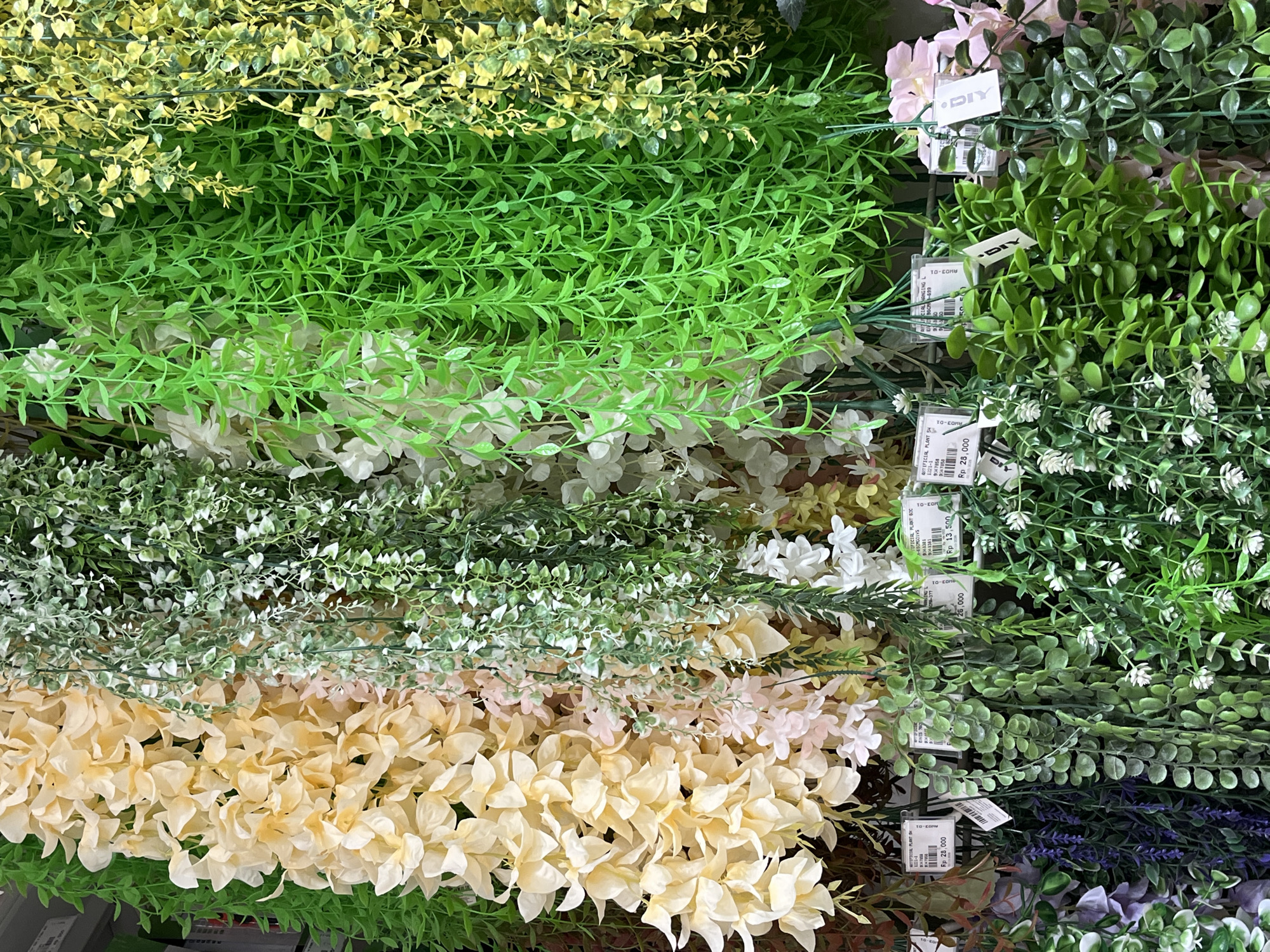 The image displays a dense retail arrangement of assorted artificial hanging plants, observed within a store located in Tangerang, Indonesia. The upper portion of the frame features vertical strands of artificial flora and foliage, prominently showcasing pale yellow wisteria-like flower clusters, interwoven light green and white trailing leaves, vibrant lime green small-leafed vines, and variegated green and yellow foliage. Descending into the lower half, additional plant varieties are visible, including artificial purple lavender-like sprigs, dark green round-leafed trailing plants, and lighter green vines adorned with small white flowers. Numerous white rectangular price tags are attached to the plants, indicating product names such as "ARTIFICIAL PLANT" and displaying prices in Indonesian Rupiah (Rp), with visible denominations including Rp 28,000 and Rp 13,500. Several tags bear a "DIY" brand logo. The background is largely obscured by the extensive plant display, revealing only indistinct shelving or a wall structure.